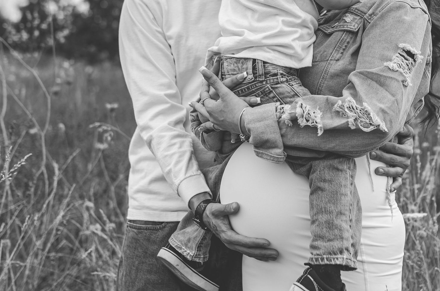 Black and white close-up of pregnant woman held by partner while carrying a child outdoors.