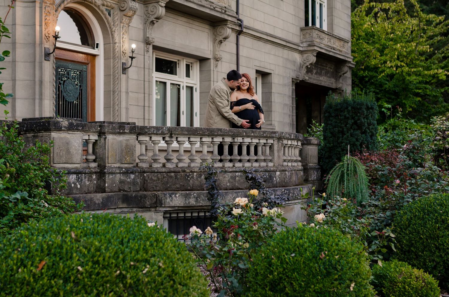 Romantic couple embracing on a stone balcony of an elegant mansion surrounded by lush gardens.