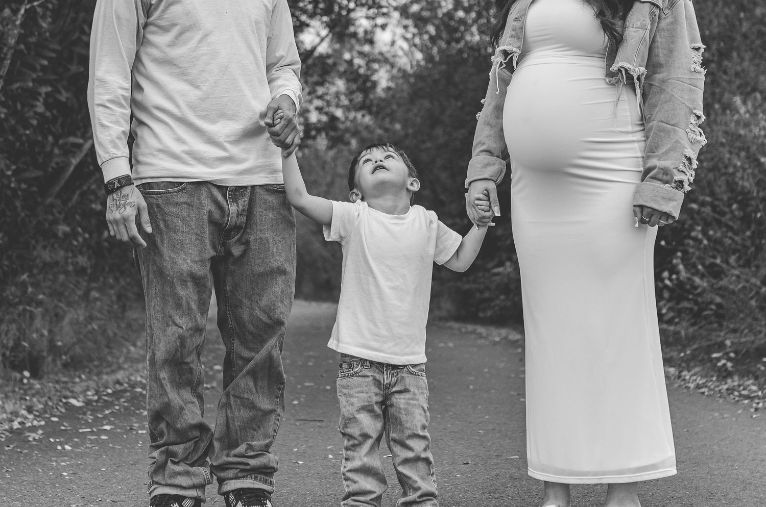 Black and white maternity photo of couple holding hands with toddler on a tree-lined path.
