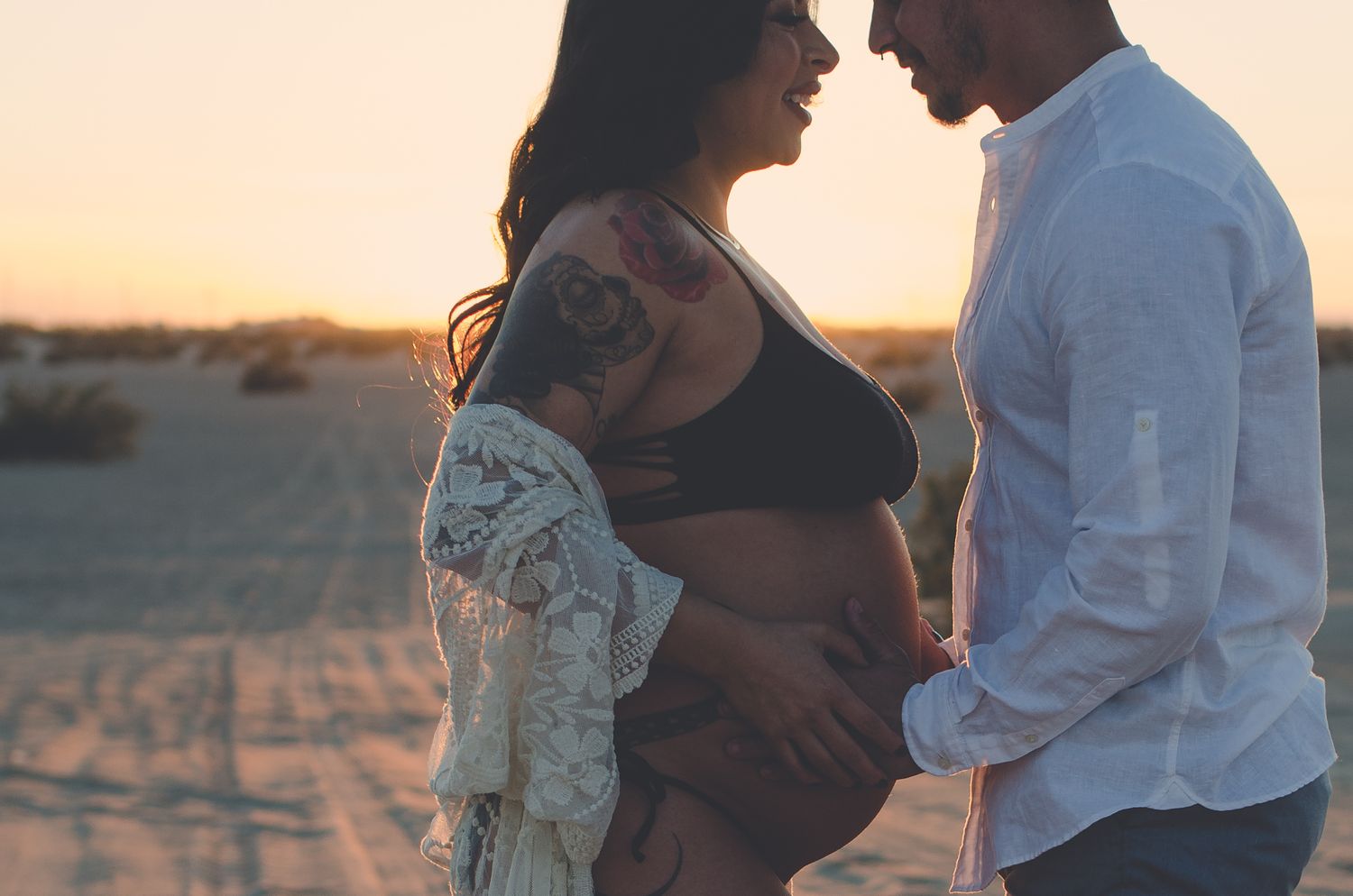 Couple sharing intimate moment at sunset on sandy desert, woman with floral tattoo showing baby bump.