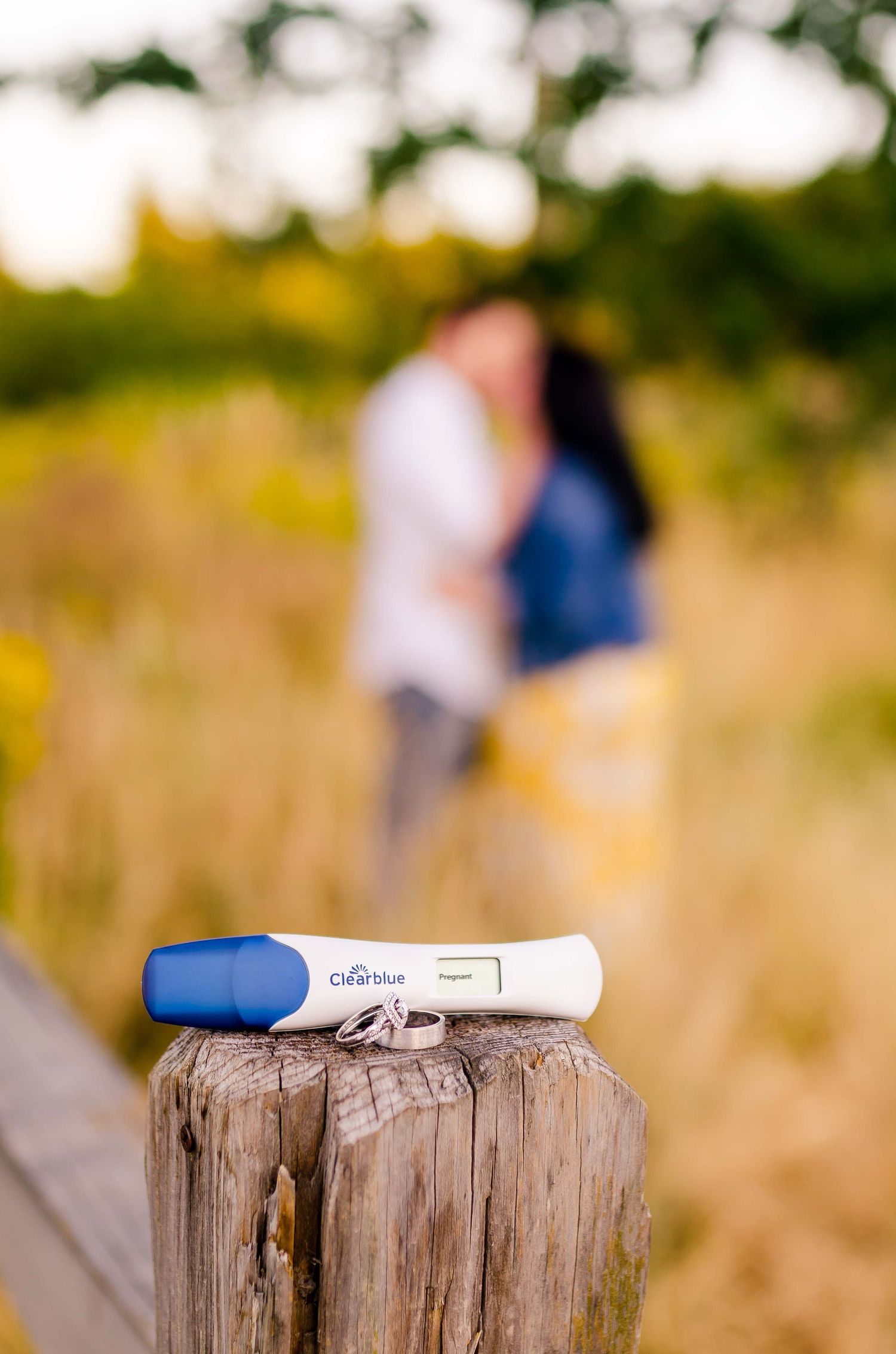 Positive Clearblue pregnancy test on wooden post with couple kissing in blurred background.