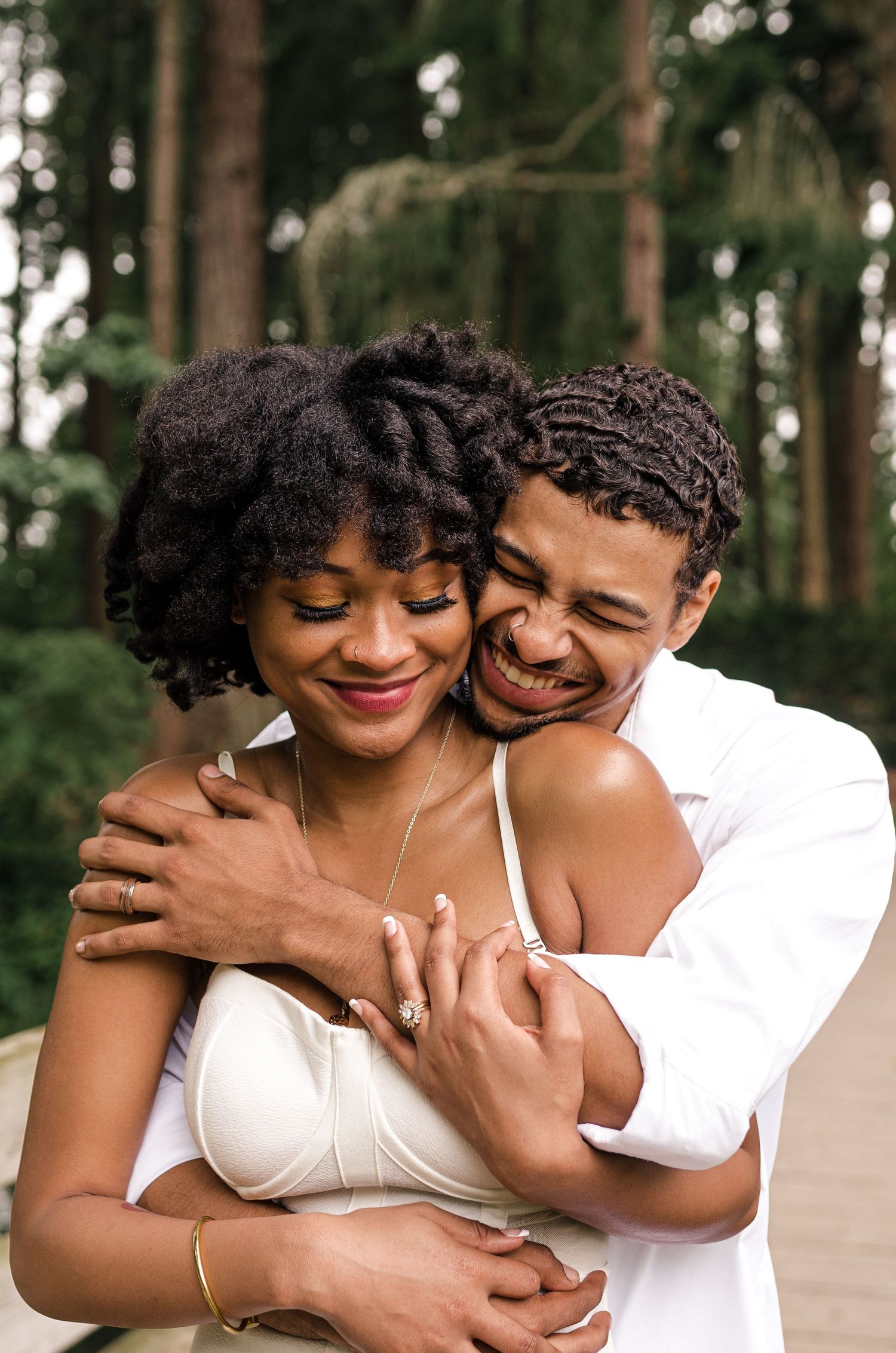 A joyful couple in white attire shares a warm embrace outdoors, surrounded by lush green trees.