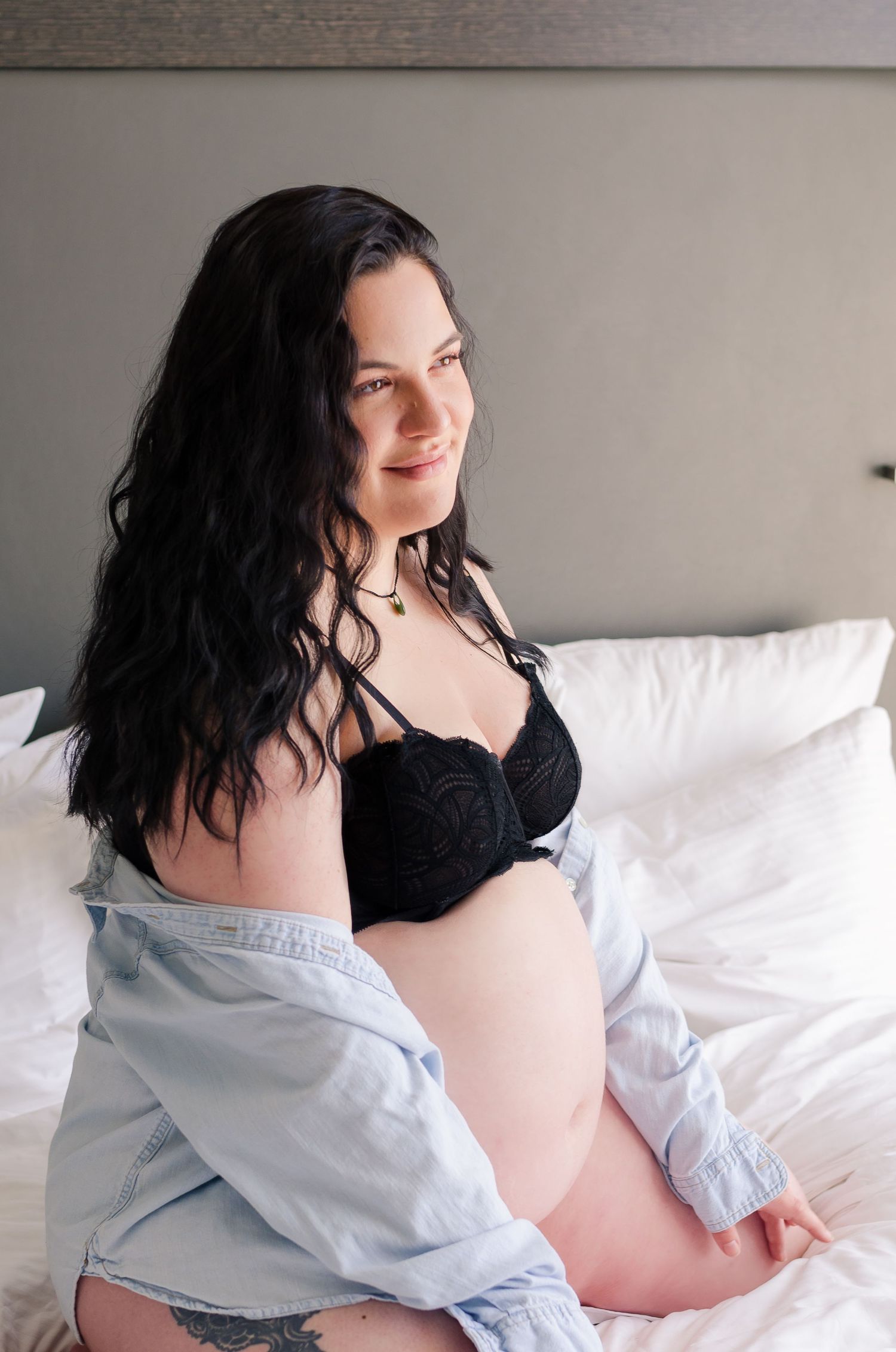 Pregnant woman with dark hair sitting on white bed wearing black lace bra and open blue shirt, smiling softly.