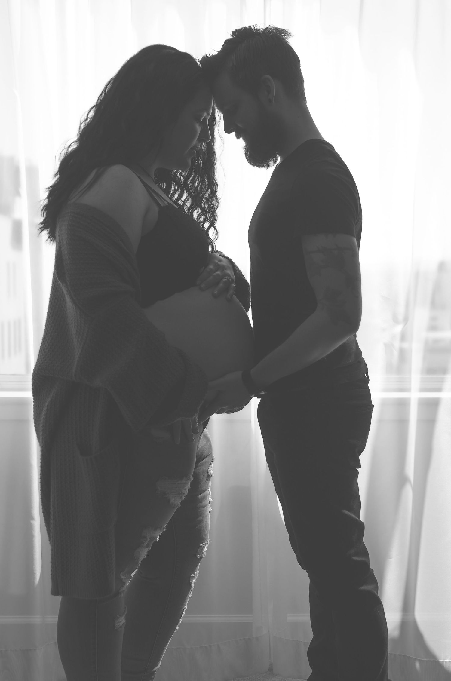 Silhouette of expectant couple touching foreheads while cradling baby bump by bright window.
