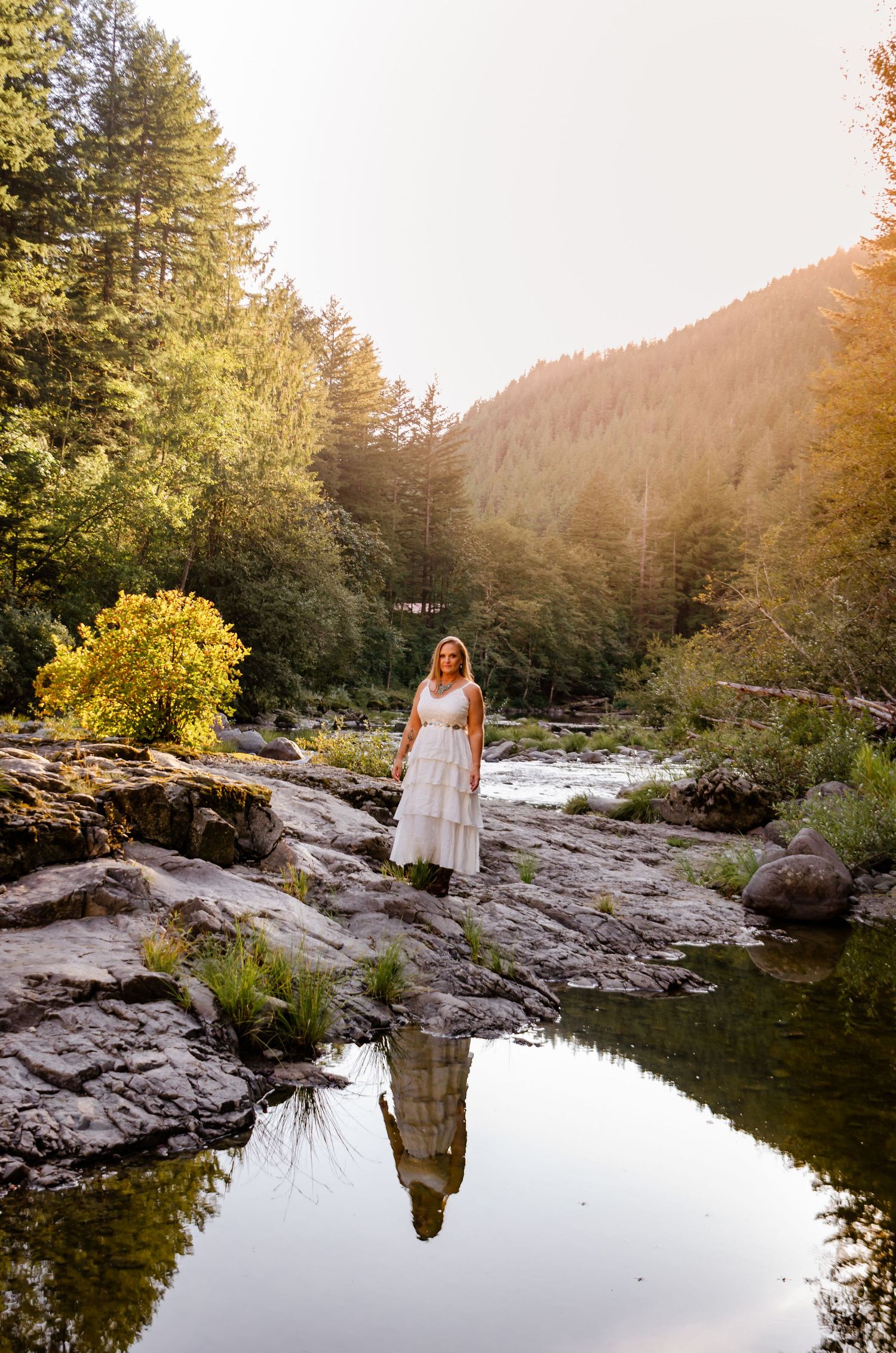 Woman in a white dress standing on rocks reflected in a calm river surrounded by forest.