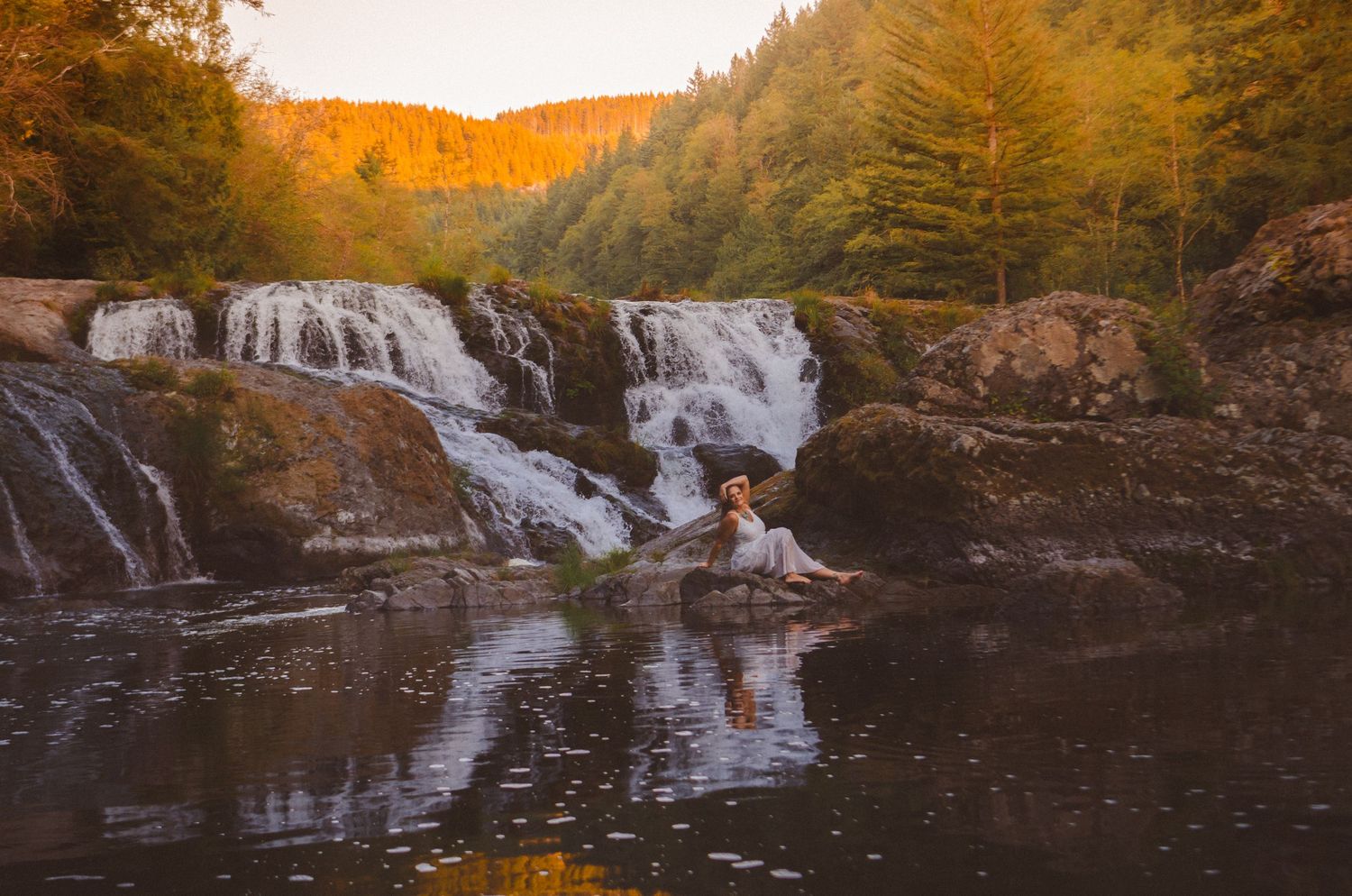 Woman in white dress sitting on rocks before a stunning autumn waterfall in the forest.