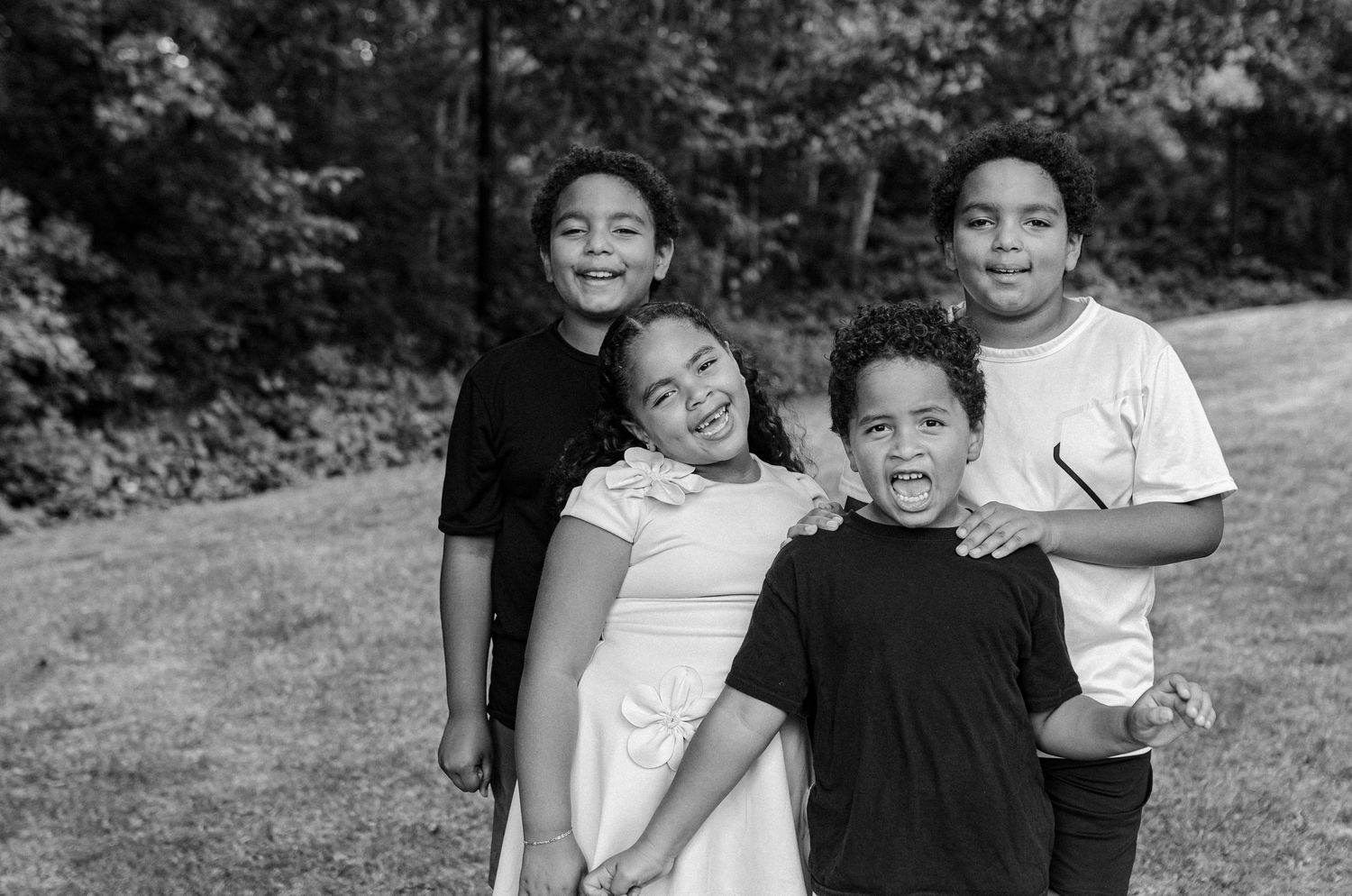 Black and white portrait of four smiling children standing together on grass with trees in background.