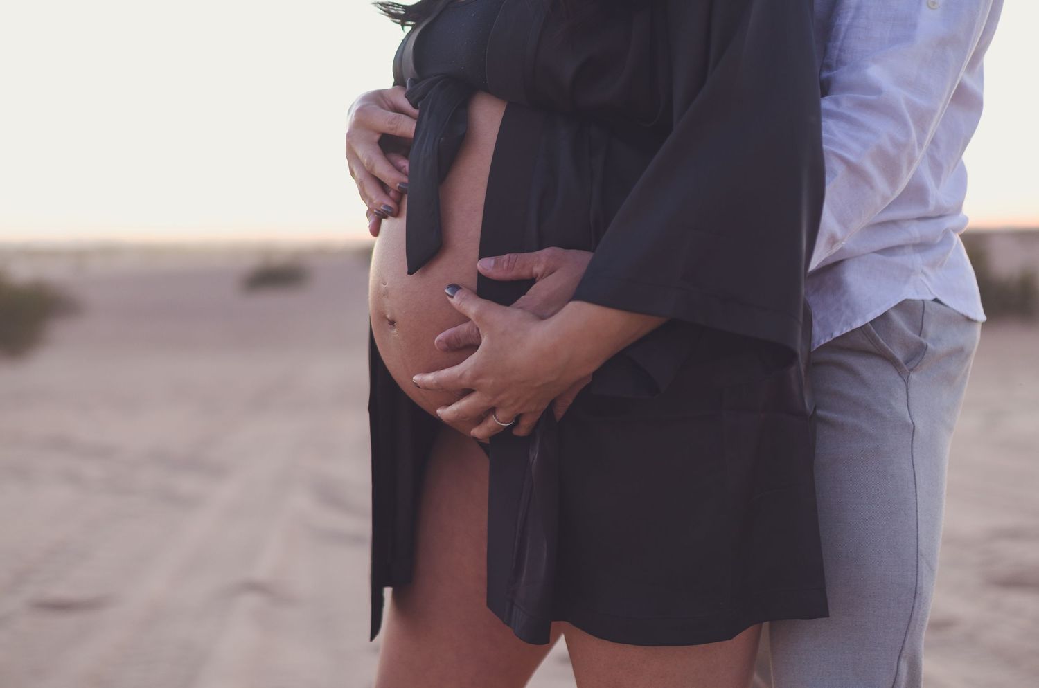 Couple tenderly cradling pregnant belly together outdoors on sandy desert at dusk.