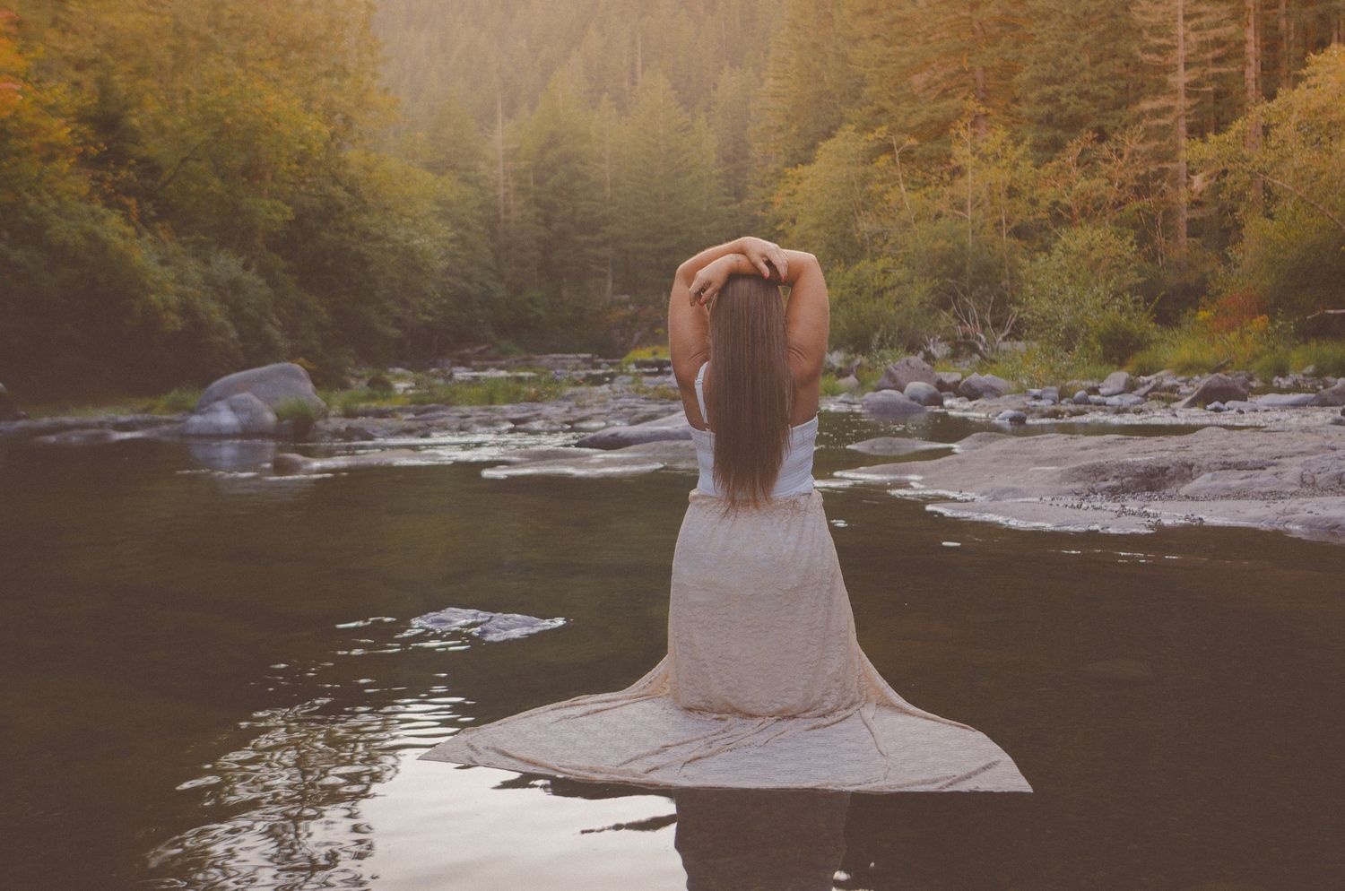 Woman in white dress sitting on rocks by a serene forest river at golden hour.
