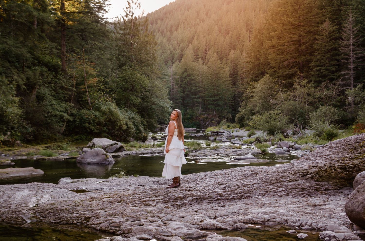Woman in a flowing white dress standing among rocky riverbed in a lush Pacific Northwest forest.