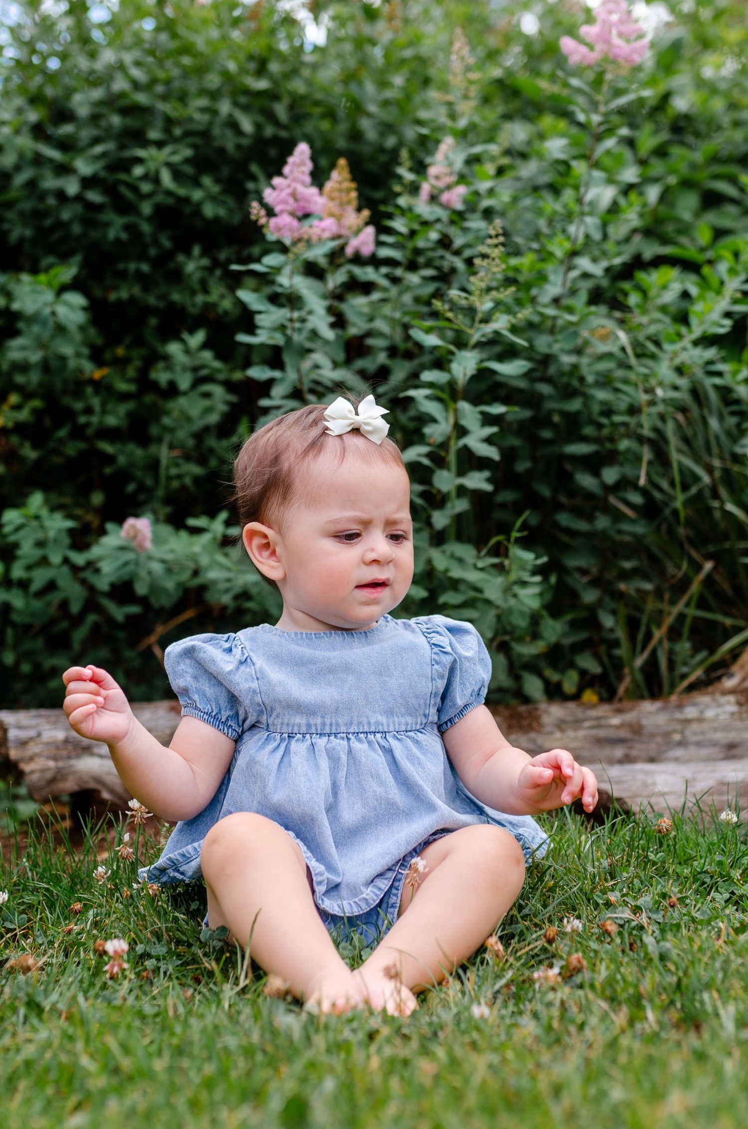 Cute baby girl wearing blue dress and white bow clip sitting cross-legged on grass in garden setting.