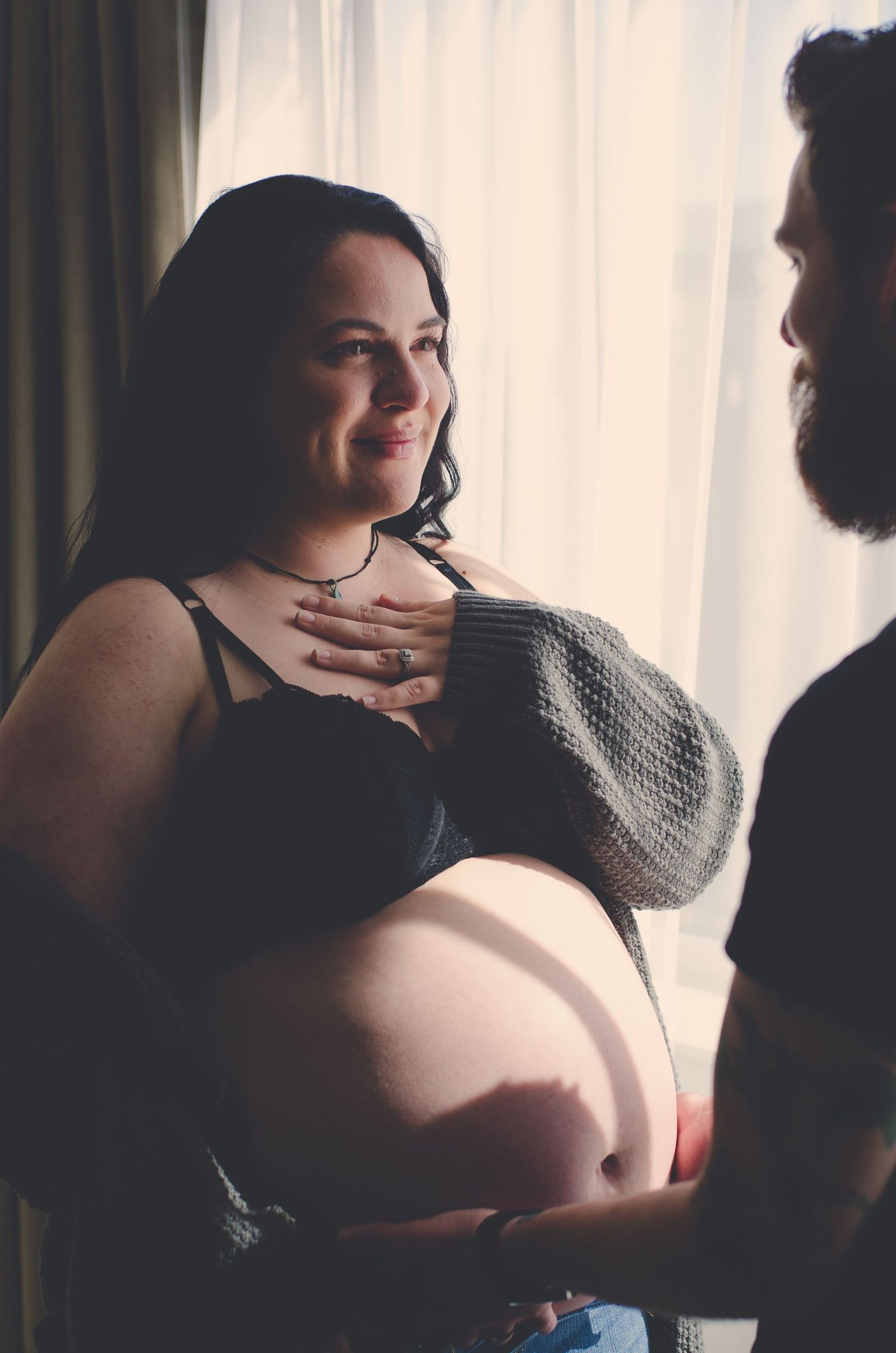 Smiling pregnant woman in black bra with partner gently touching her chest by window.