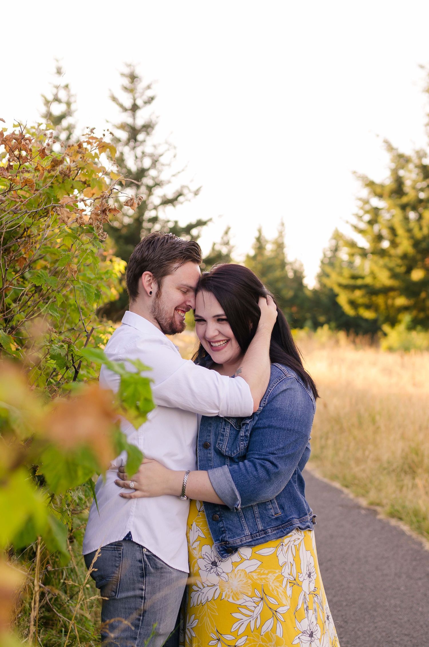 Couple embracing and smiling together on a sunny path surrounded by green trees and golden grass.