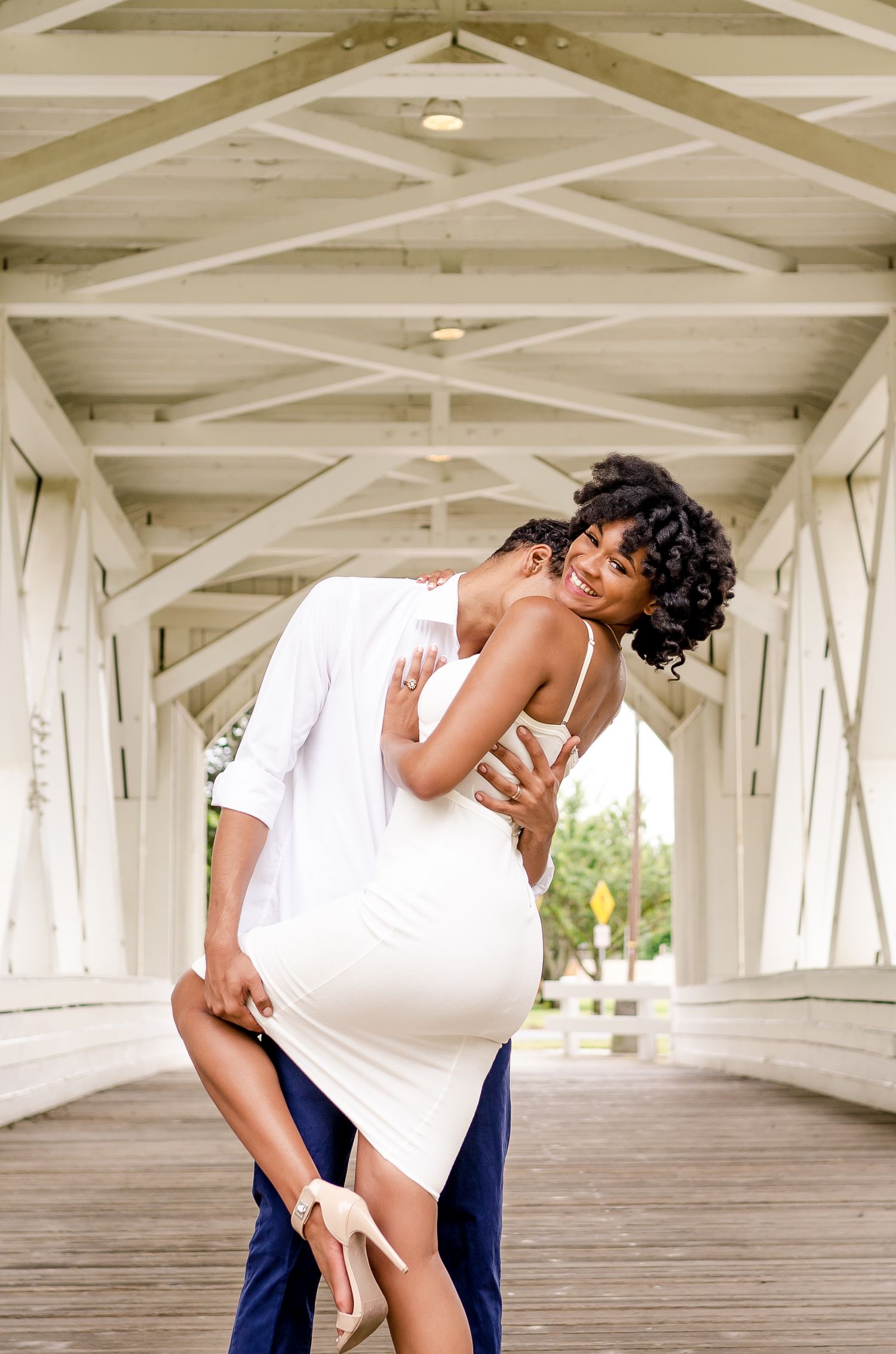 A couple shares a romantic embrace on a white wooden covered bridge, woman in white dress lifted by her partner.