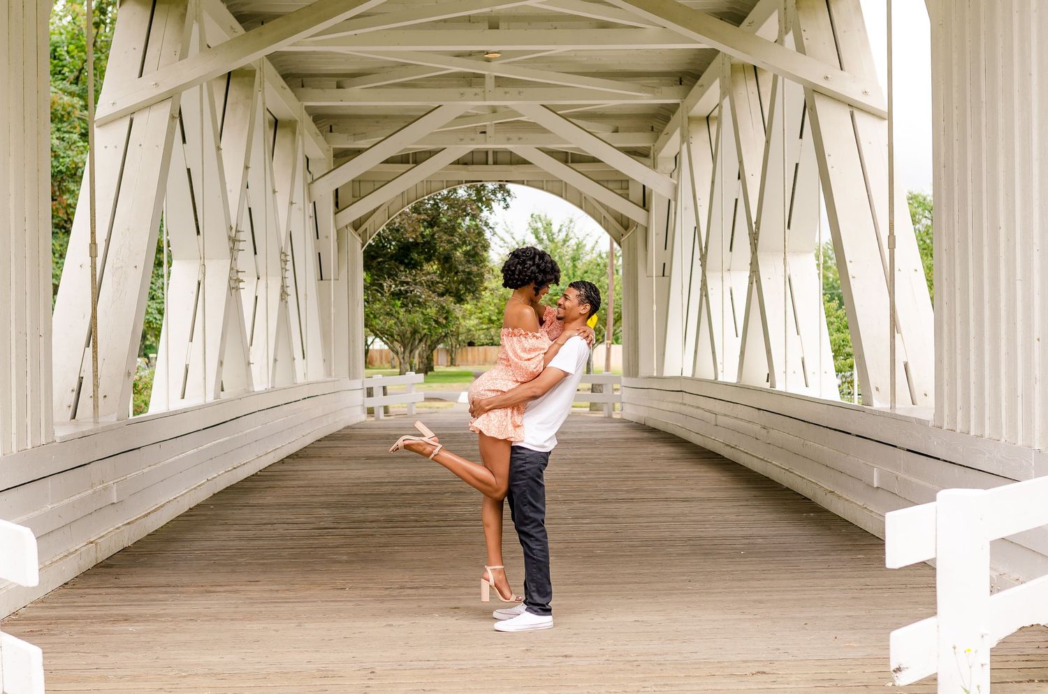 Couple sharing a romantic lift embrace inside a white wooden covered bridge during an engagement photo session.
