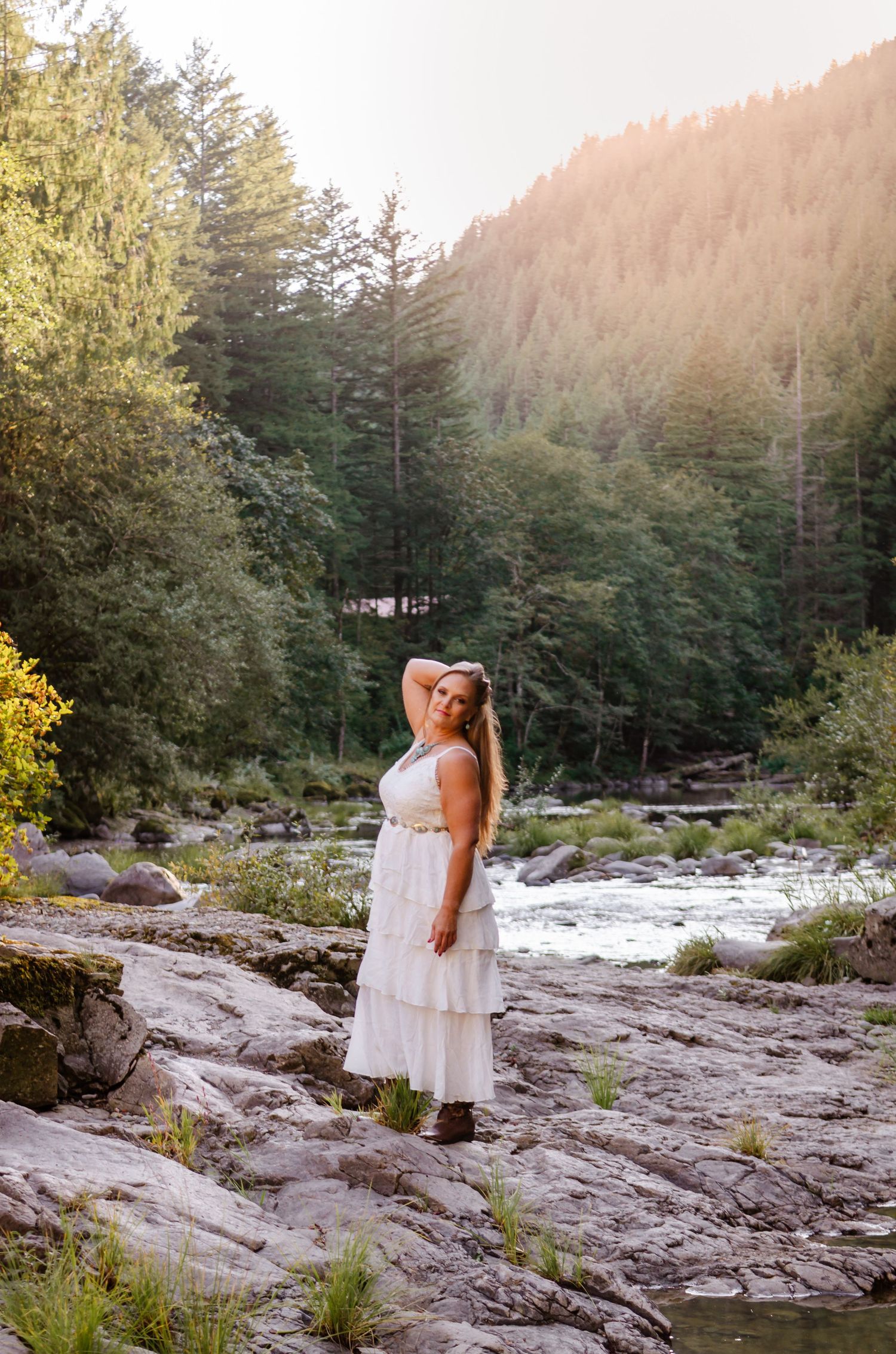 Woman in white tiered dress posing by a rocky mountain river surrounded by tall pine trees.