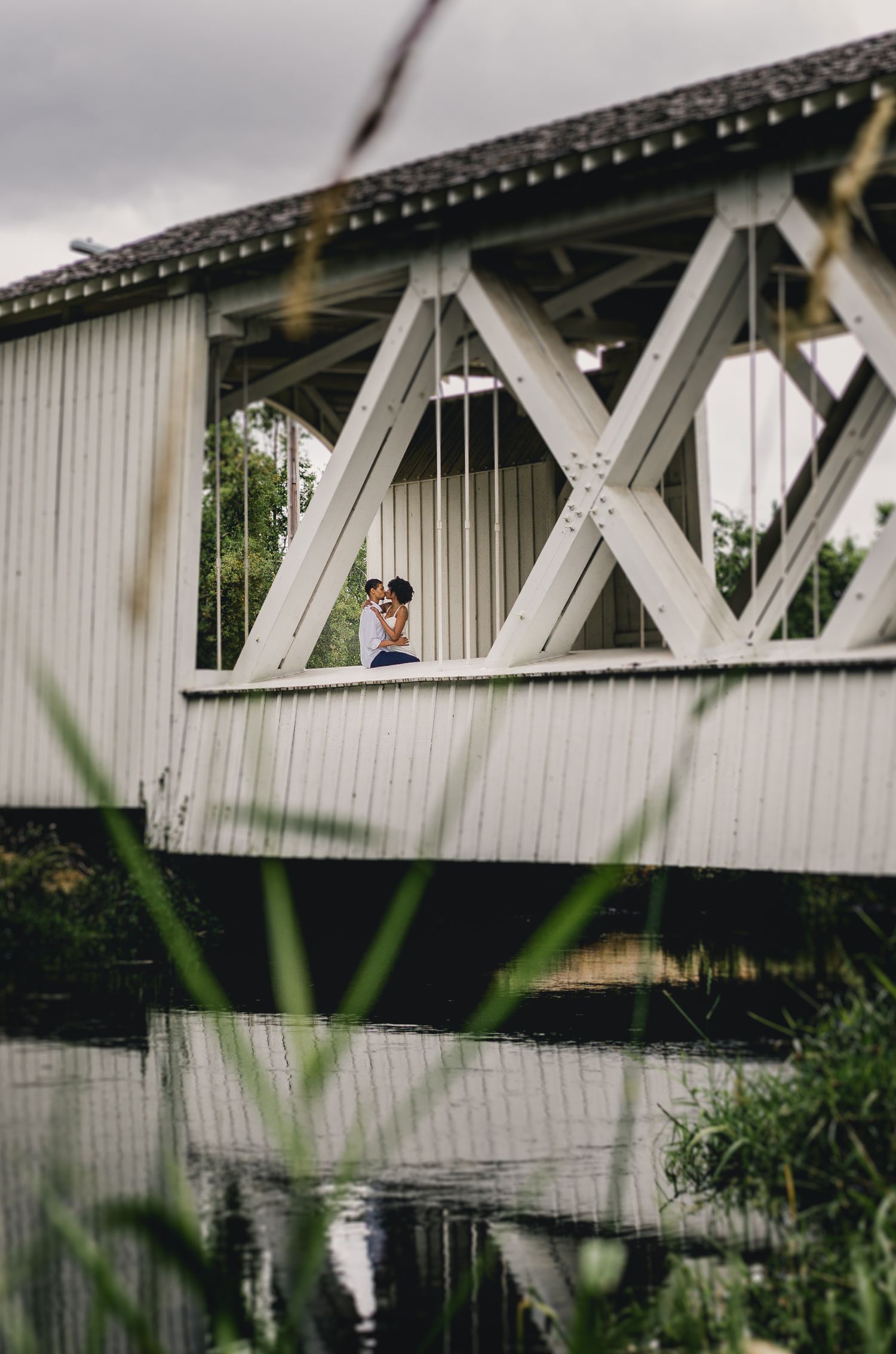 Couple sharing a romantic embrace inside a white covered bridge reflected over calm water.