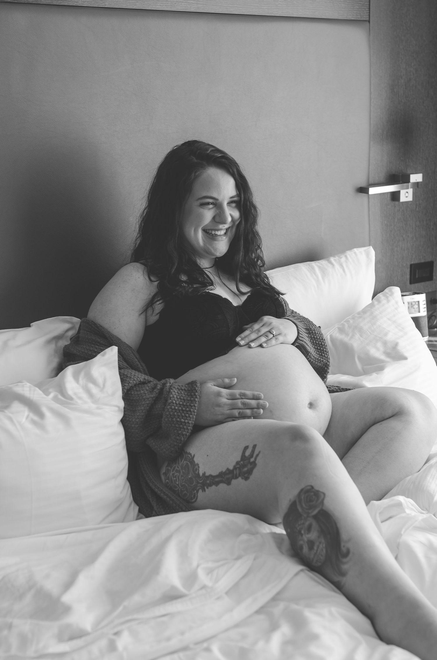 Happy pregnant woman with tattoos laughing while sitting on white bed in black lingerie.