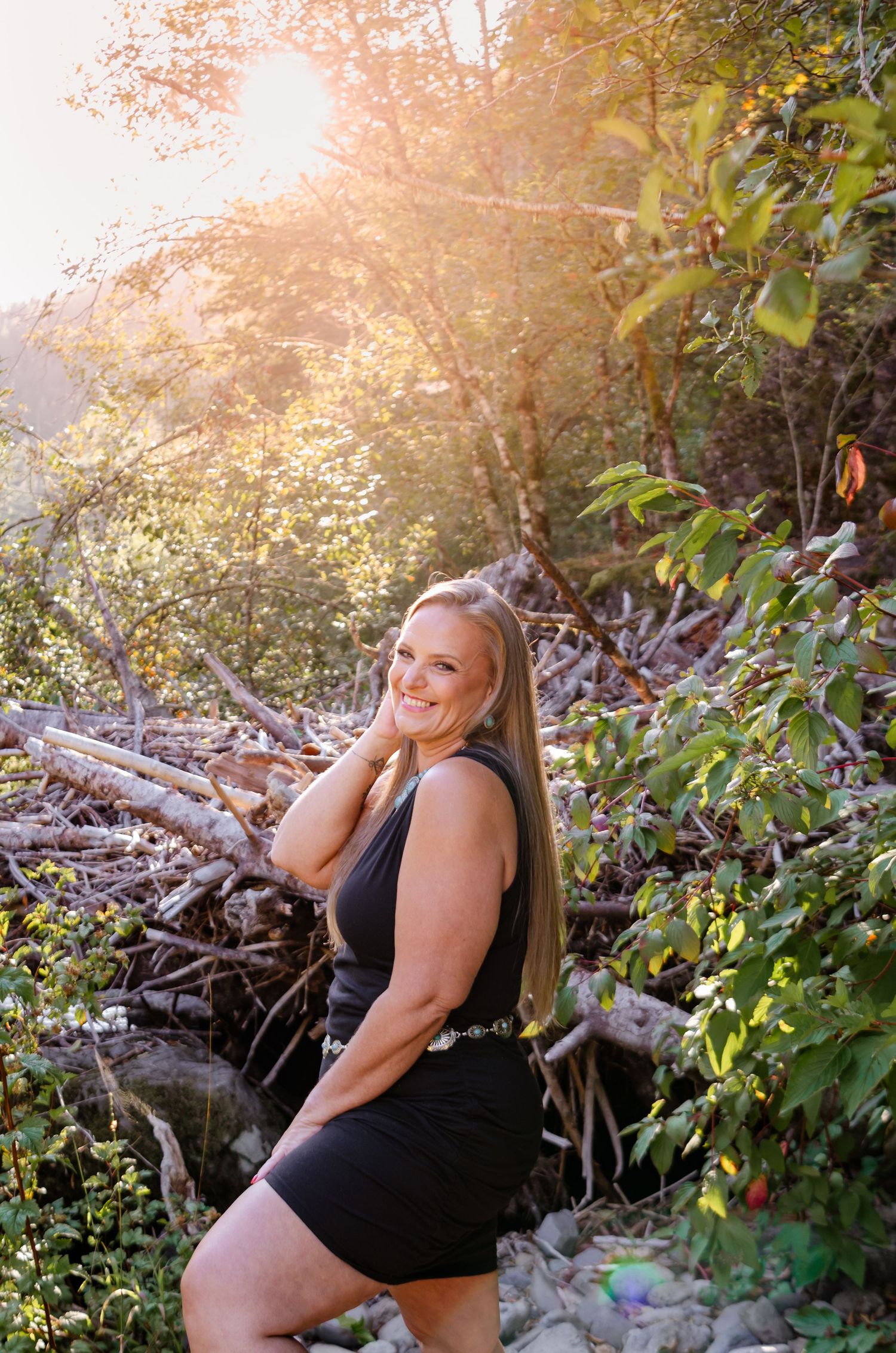 Smiling blonde woman in a black romper posing in a sunlit forest with golden light.