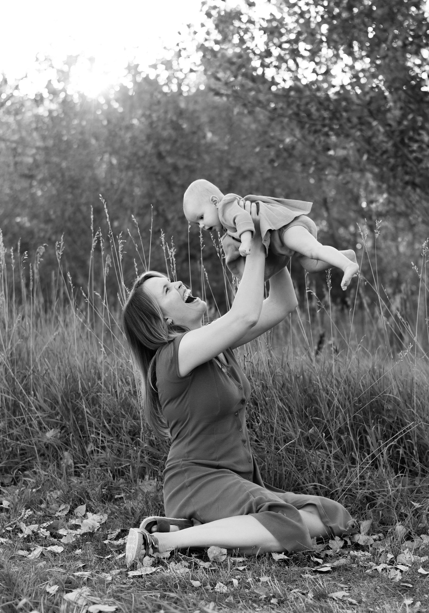 A tender black and white moment captured in tall grass as a baby is lifted joyfully into the air.