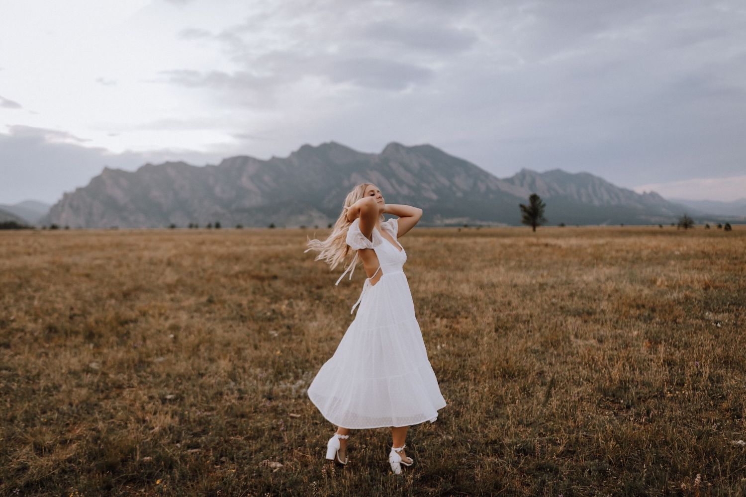 Woman in white dress twirling in field with mountains in background at sunset.