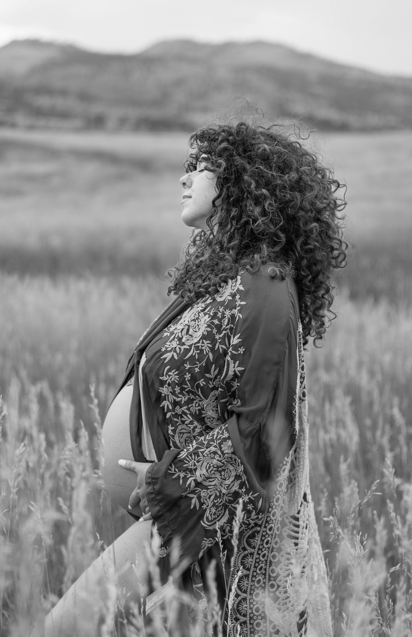 Black and white portrait of woman with curly hair in embroidered dress standing in field for maternity photos.