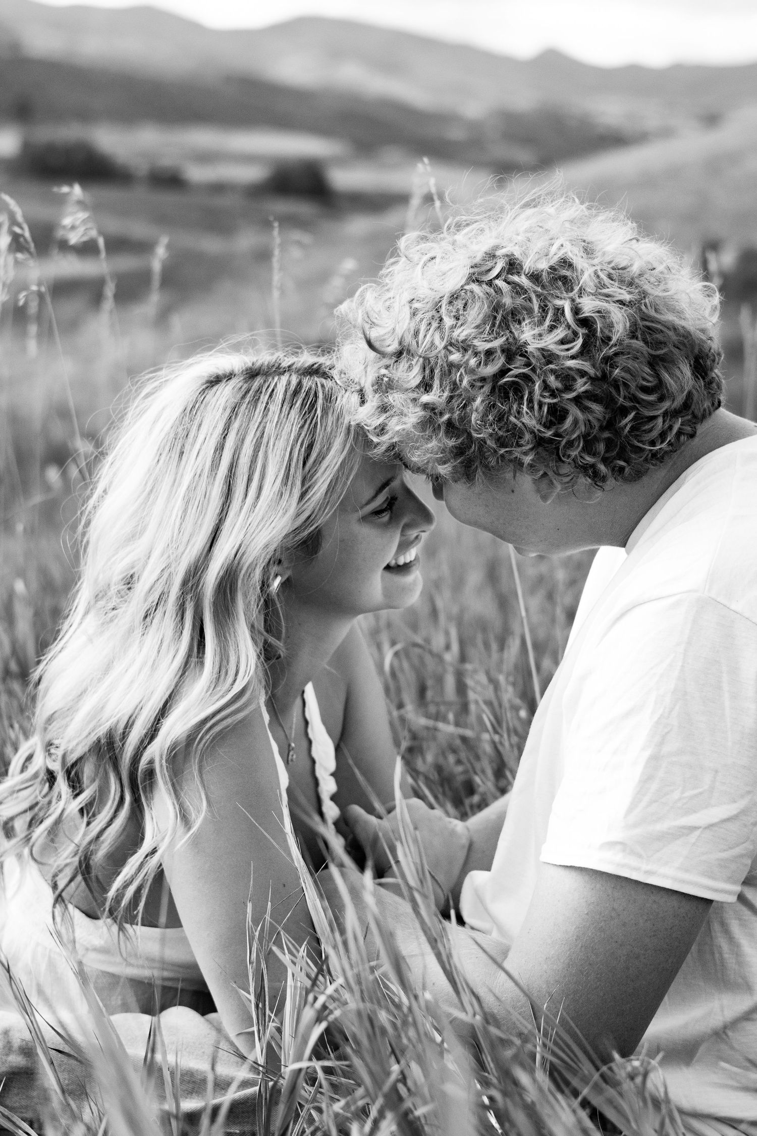 Couple shares intimate moment in field with mountains and natural lighting creating dreamy atmosphere.
