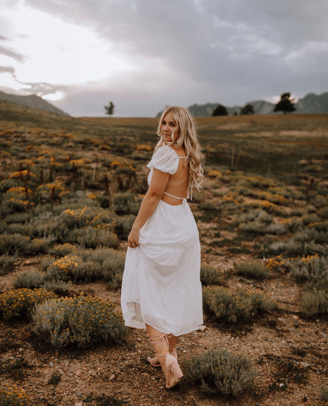 Woman in white dress walks through desert landscape with mountains and sagebrush at sunset.