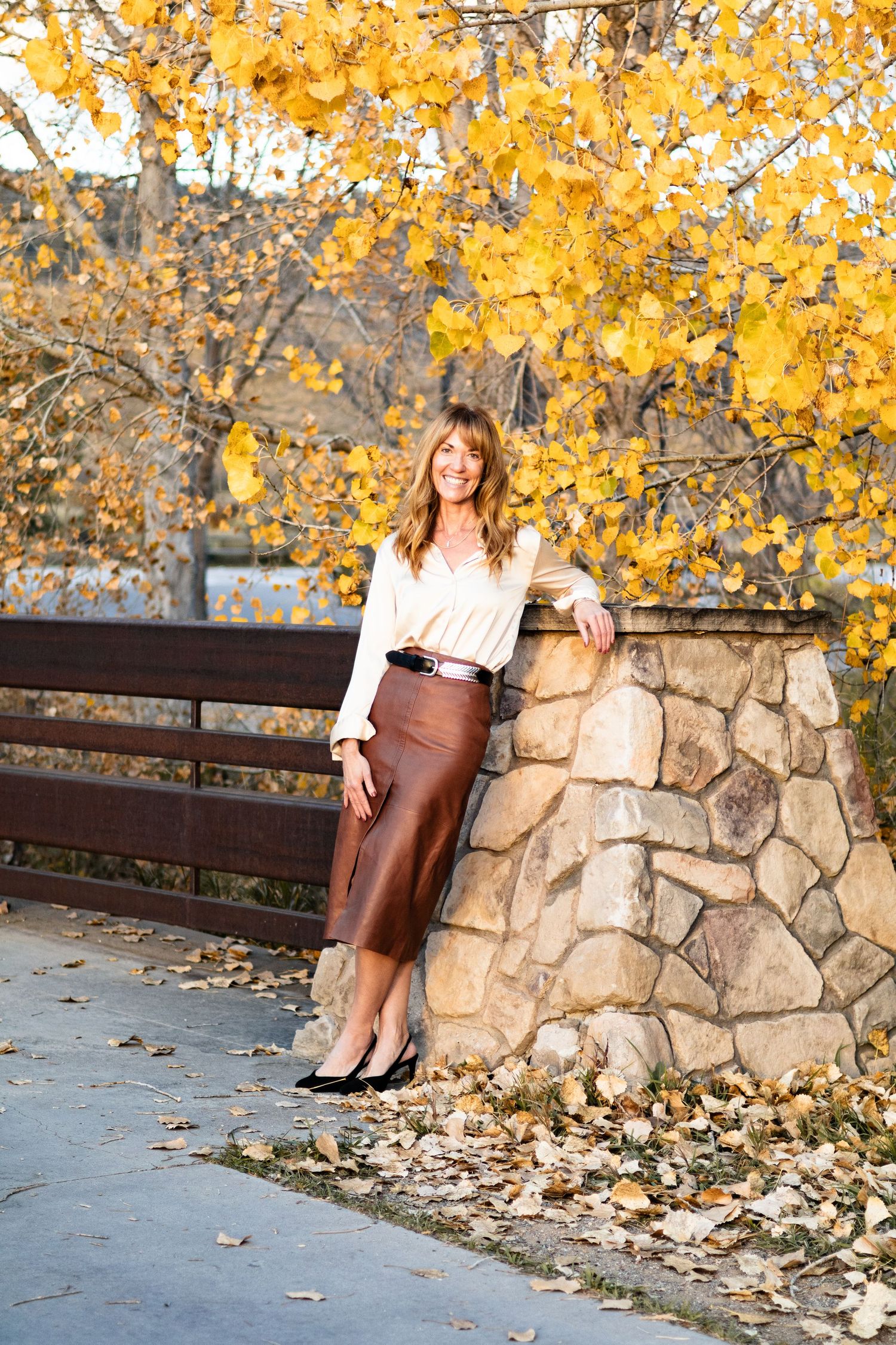 Woman in brown leather skirt poses against stone wall with golden autumn leaves in background.