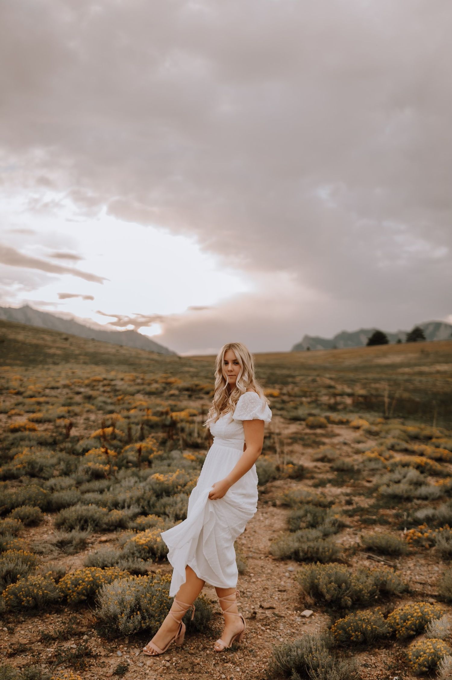 White dress and boots against a desert landscape with mountains and cloudy skies at sunset.