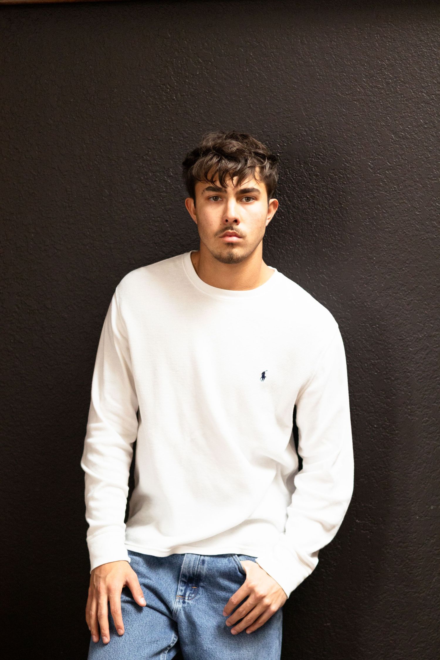 Young man in white sweater poses against dark wall in casual portrait.