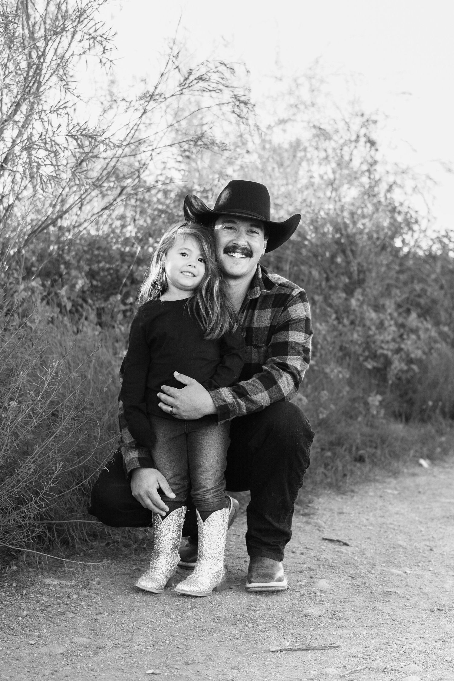 A daughter and dad in casual attire smiling for a photo on a dirt path surrounded by brush.