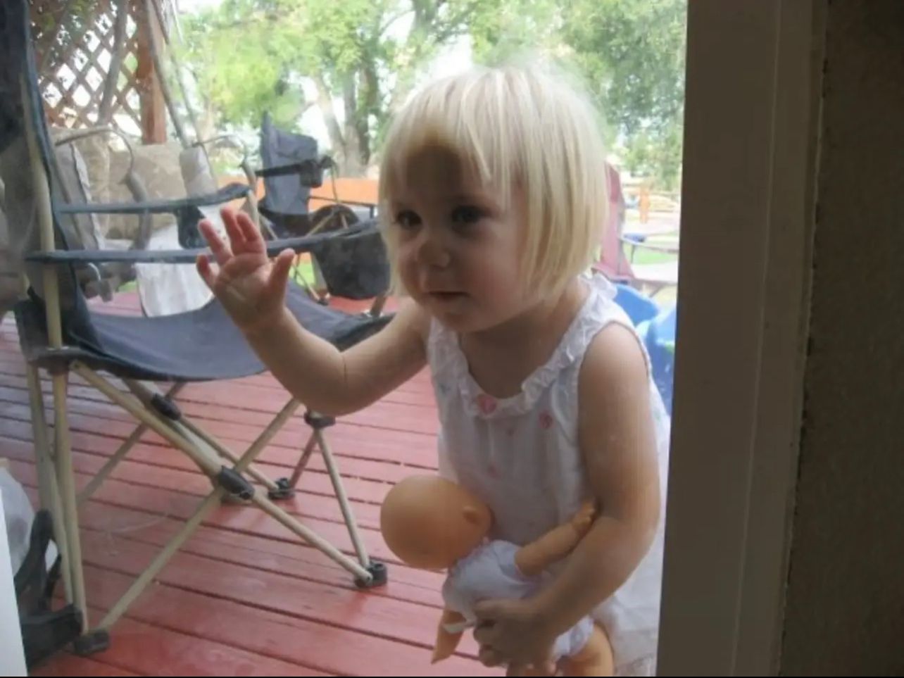 Small child in white dress reaching up while standing on a wooden deck.