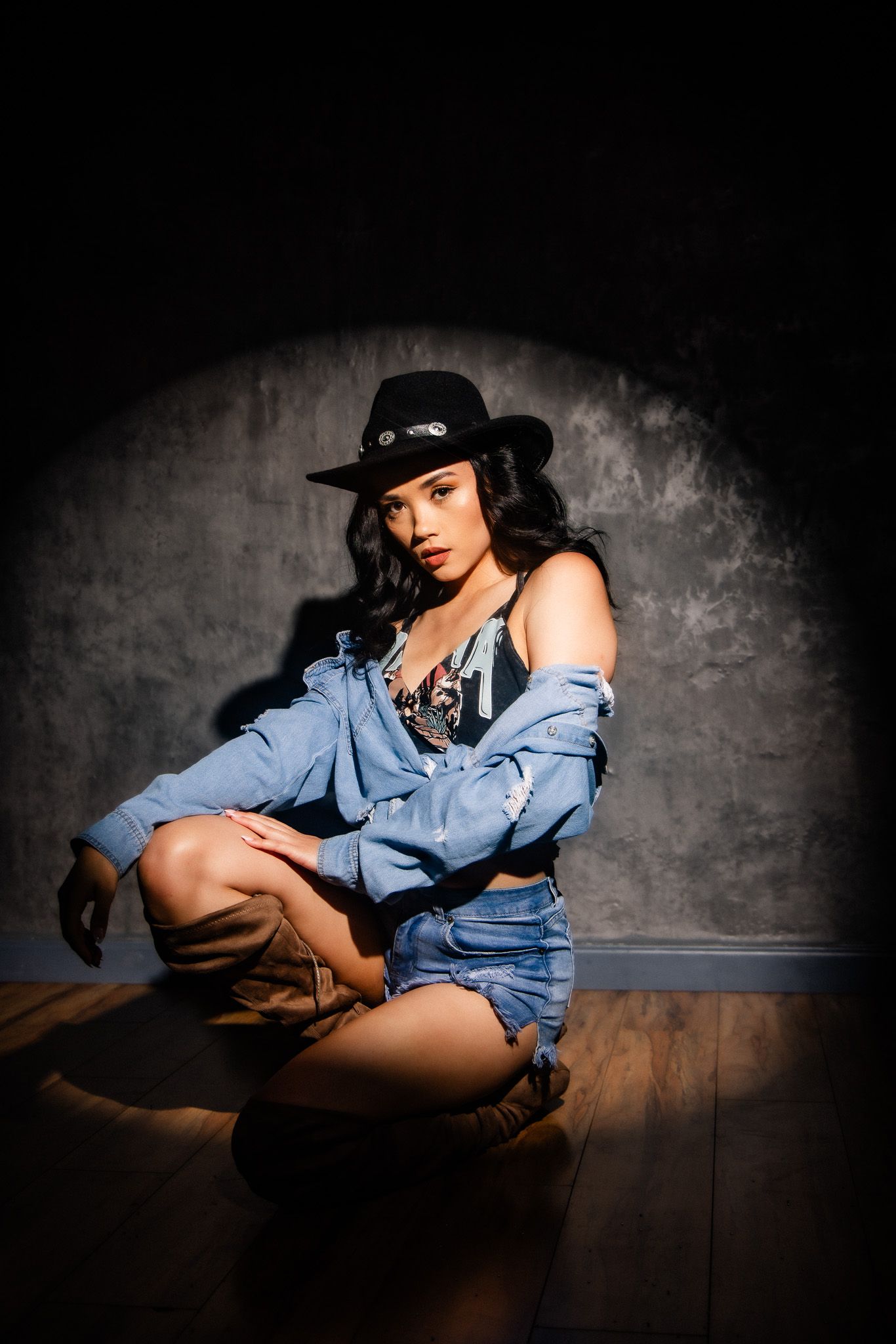 Fashion photo of model in denim shirt and hat posing dramatically against dark textured wall in low key lighting.