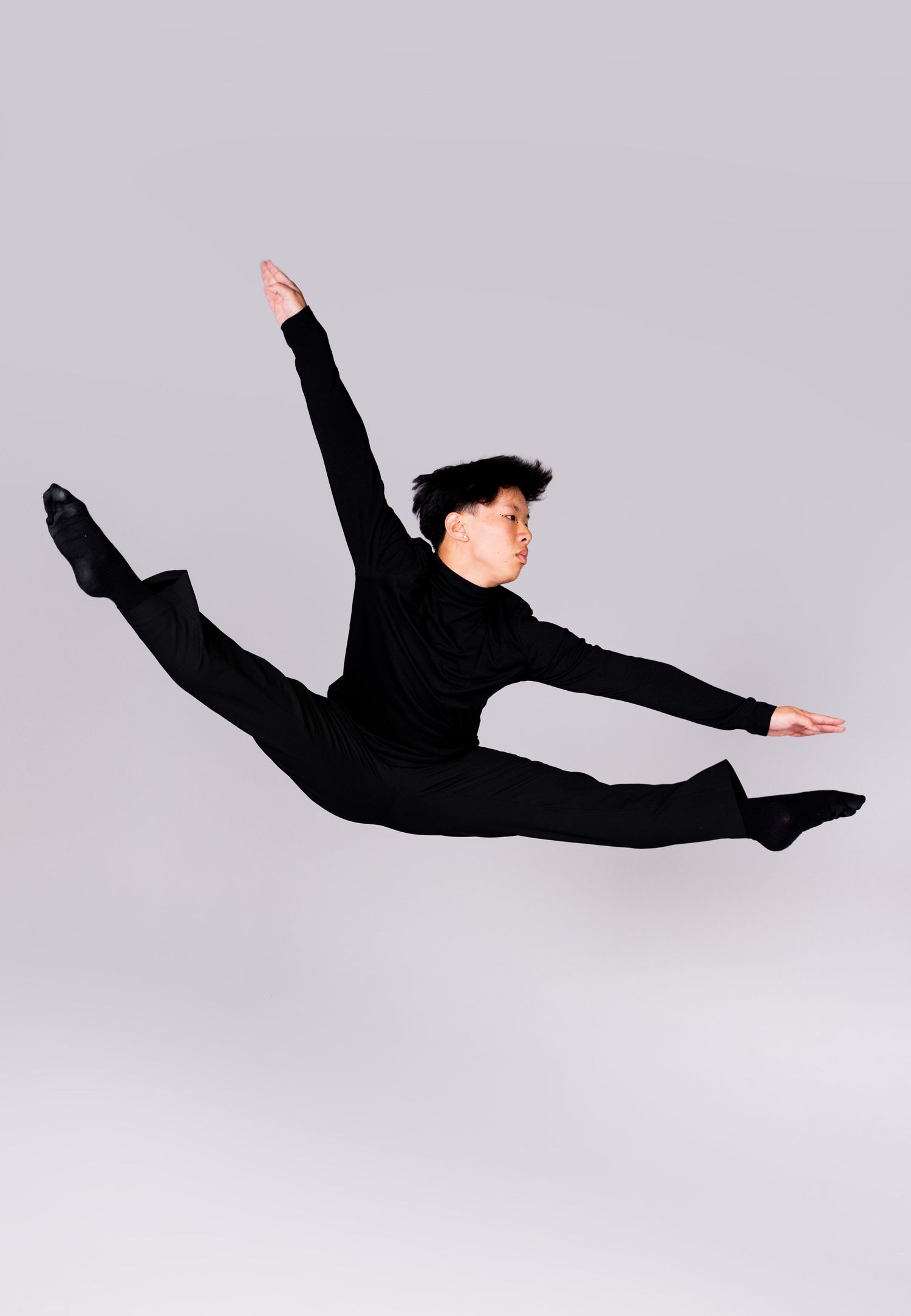 Dancer executes a graceful aerial split jump in all-black attire on gray background.