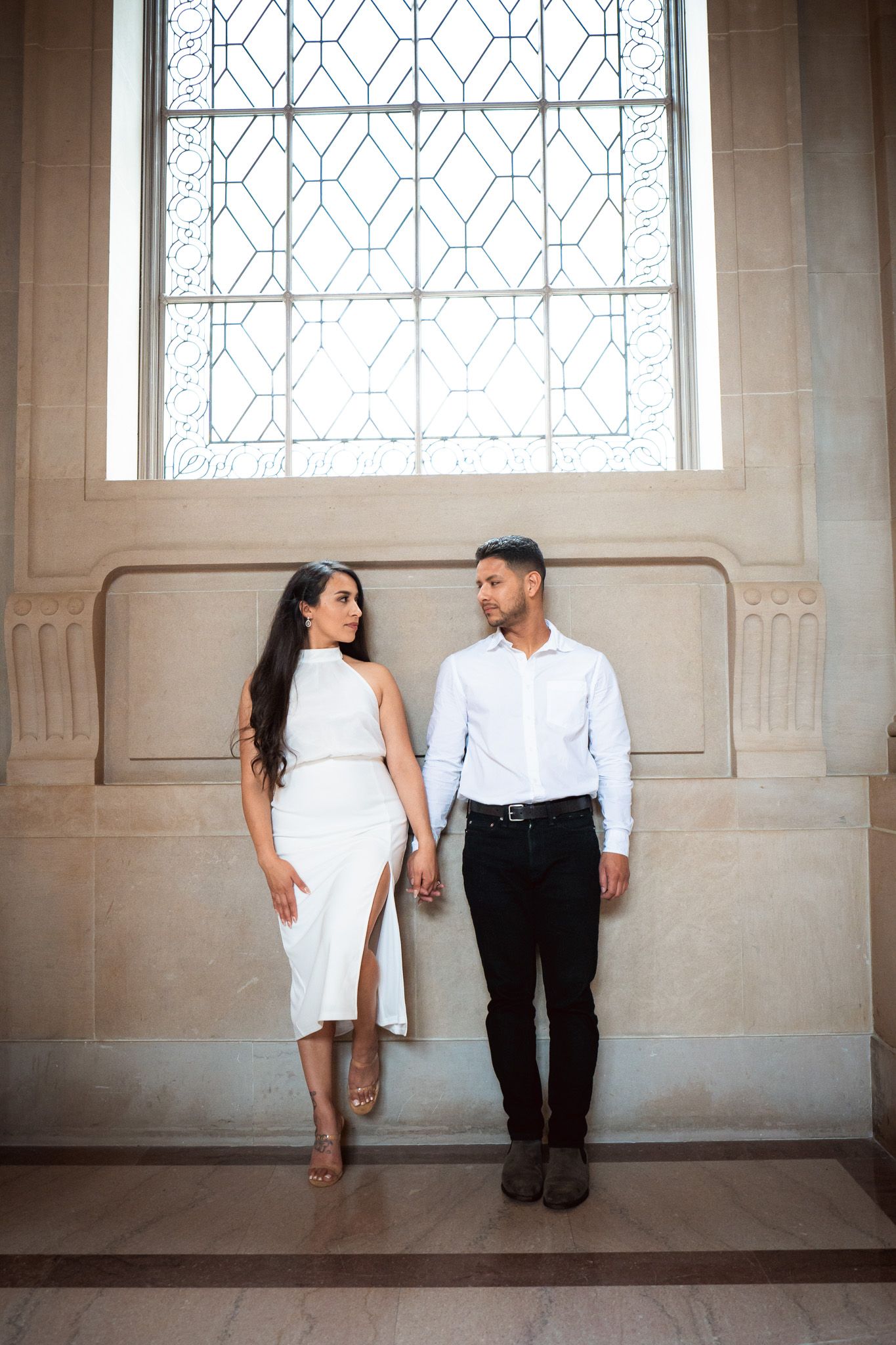 A couple in elegant attire stands together against a stone wall with a decorative window in an architectural setting.