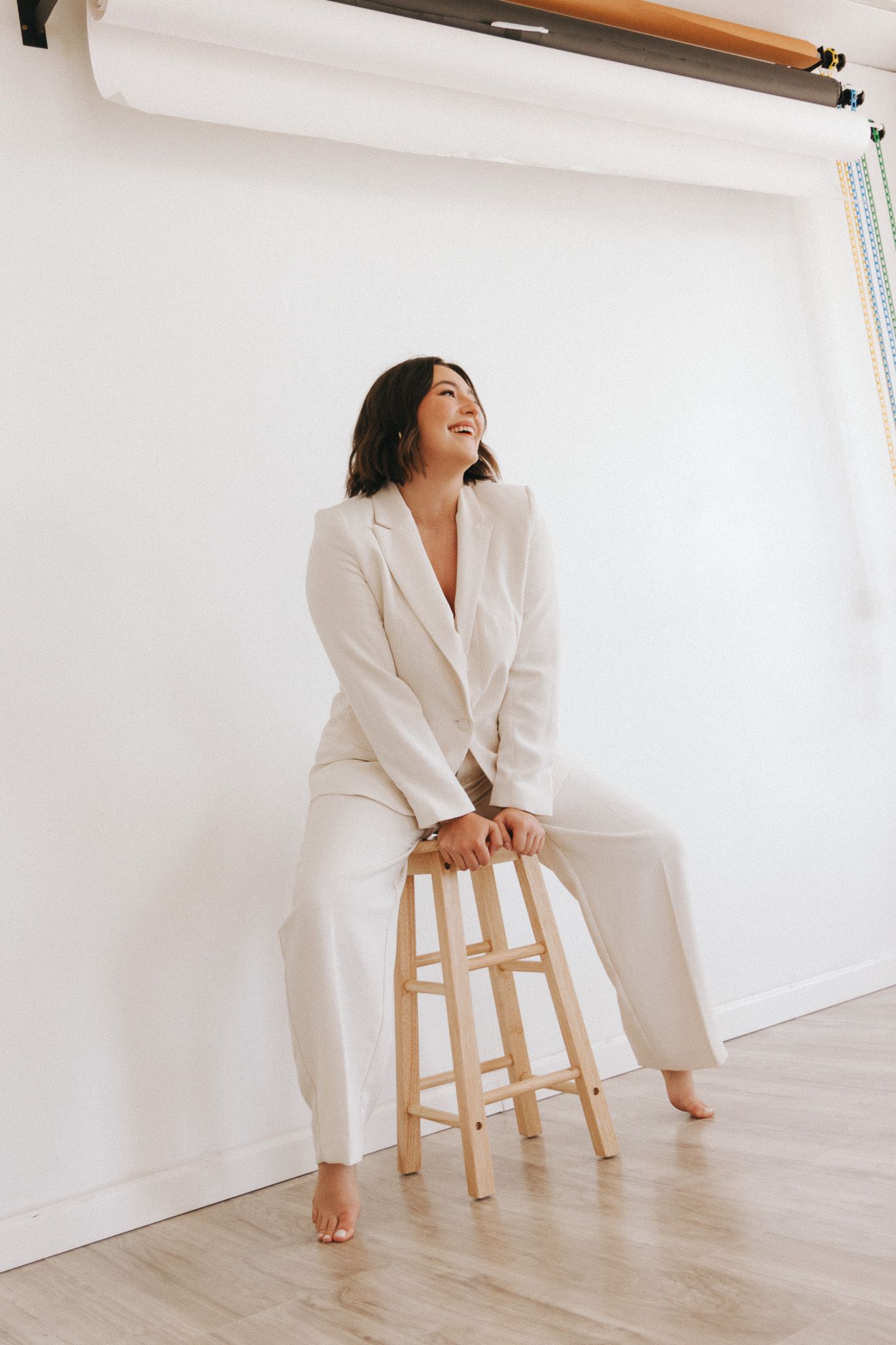 Remote worker in crisp white suit poses casually by wooden stool in bright minimalist home office setting.