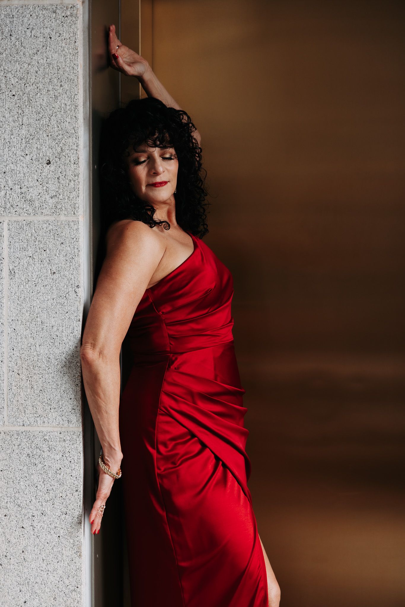 A person wearing a flowing red evening dress poses dramatically against a concrete wall in moody lighting.
