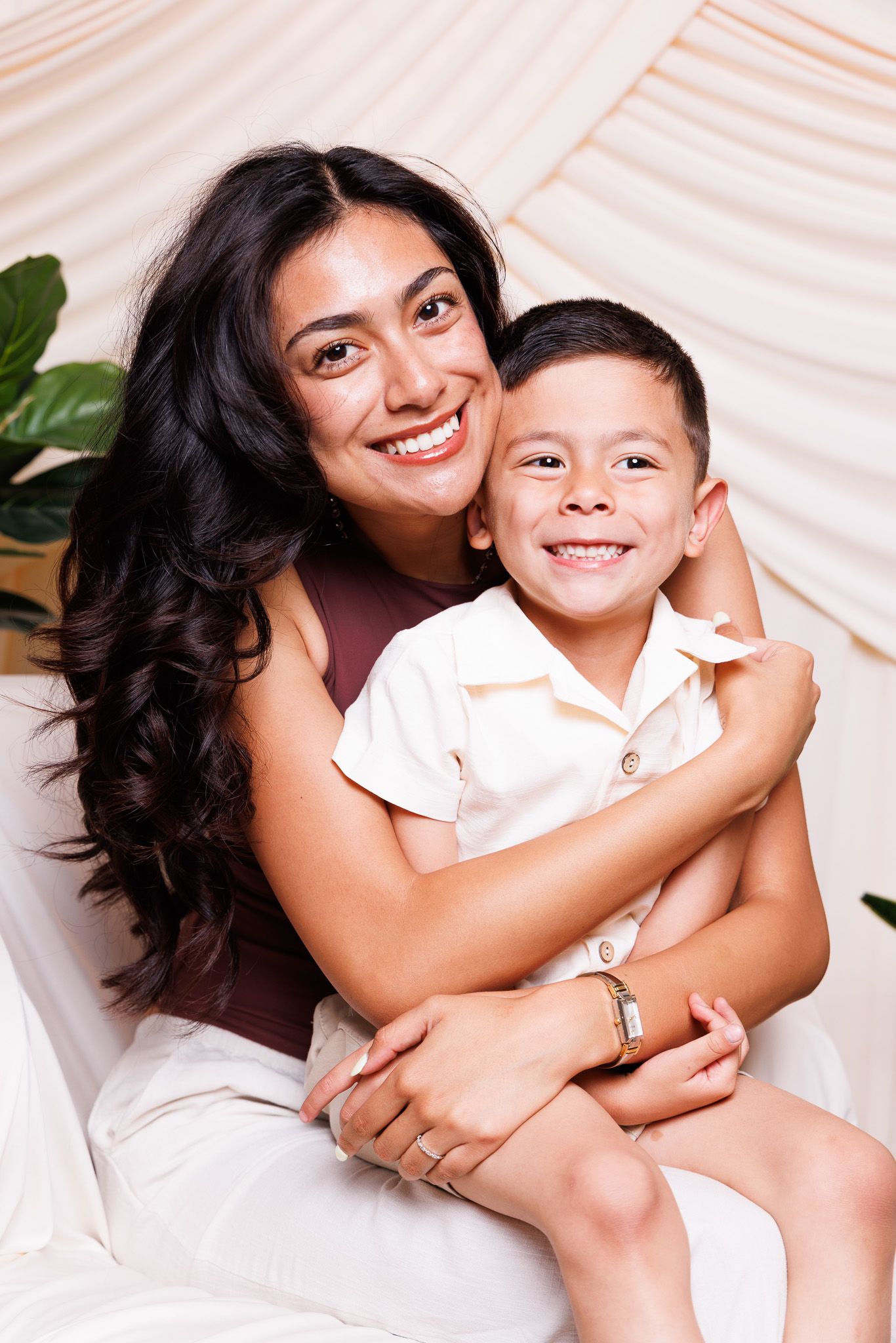 A smiling woman with long dark hair hugs a young boy in a cream shirt against a soft beige backdrop.