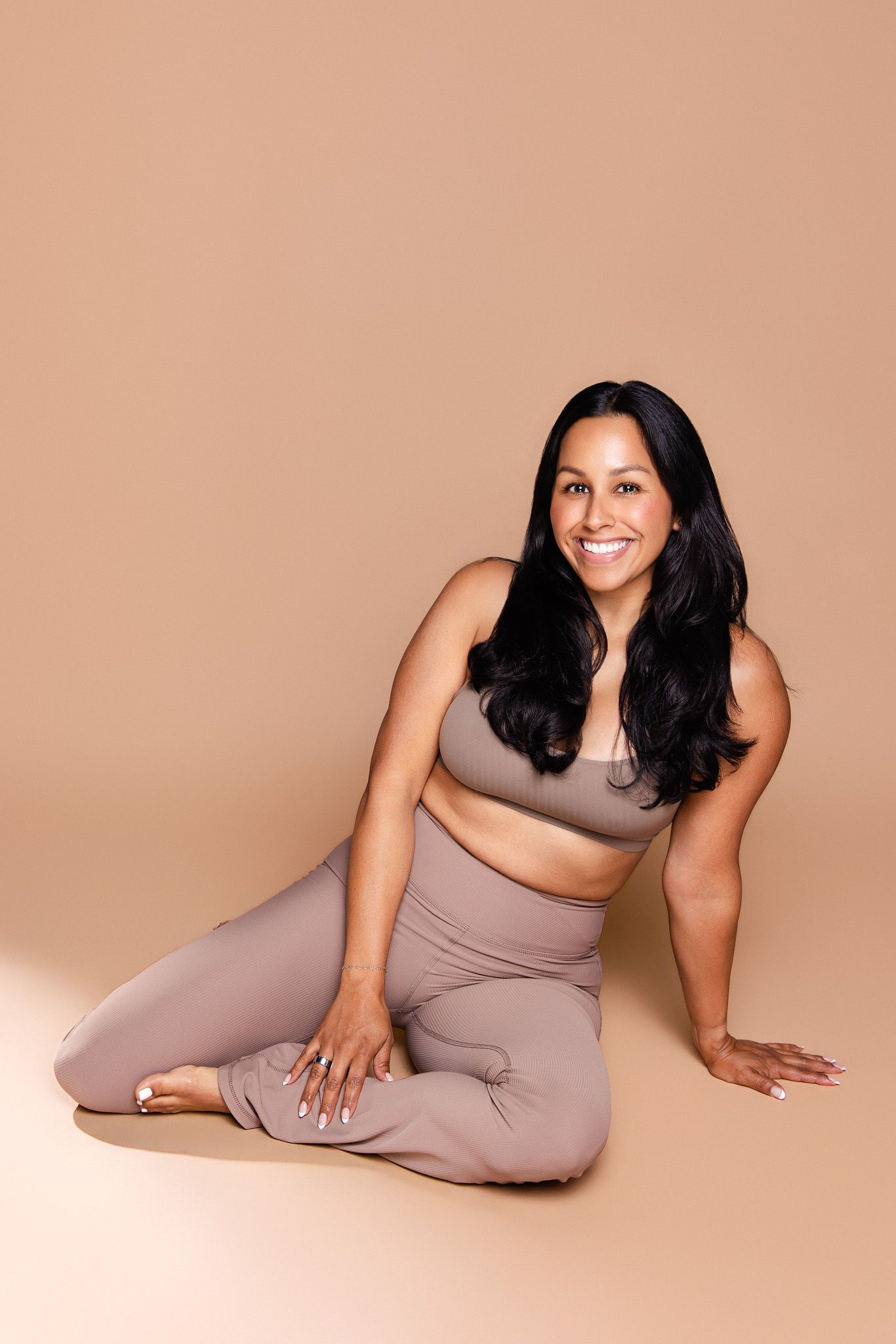 Smiling woman in taupe activewear set sitting gracefully on beige studio floor.