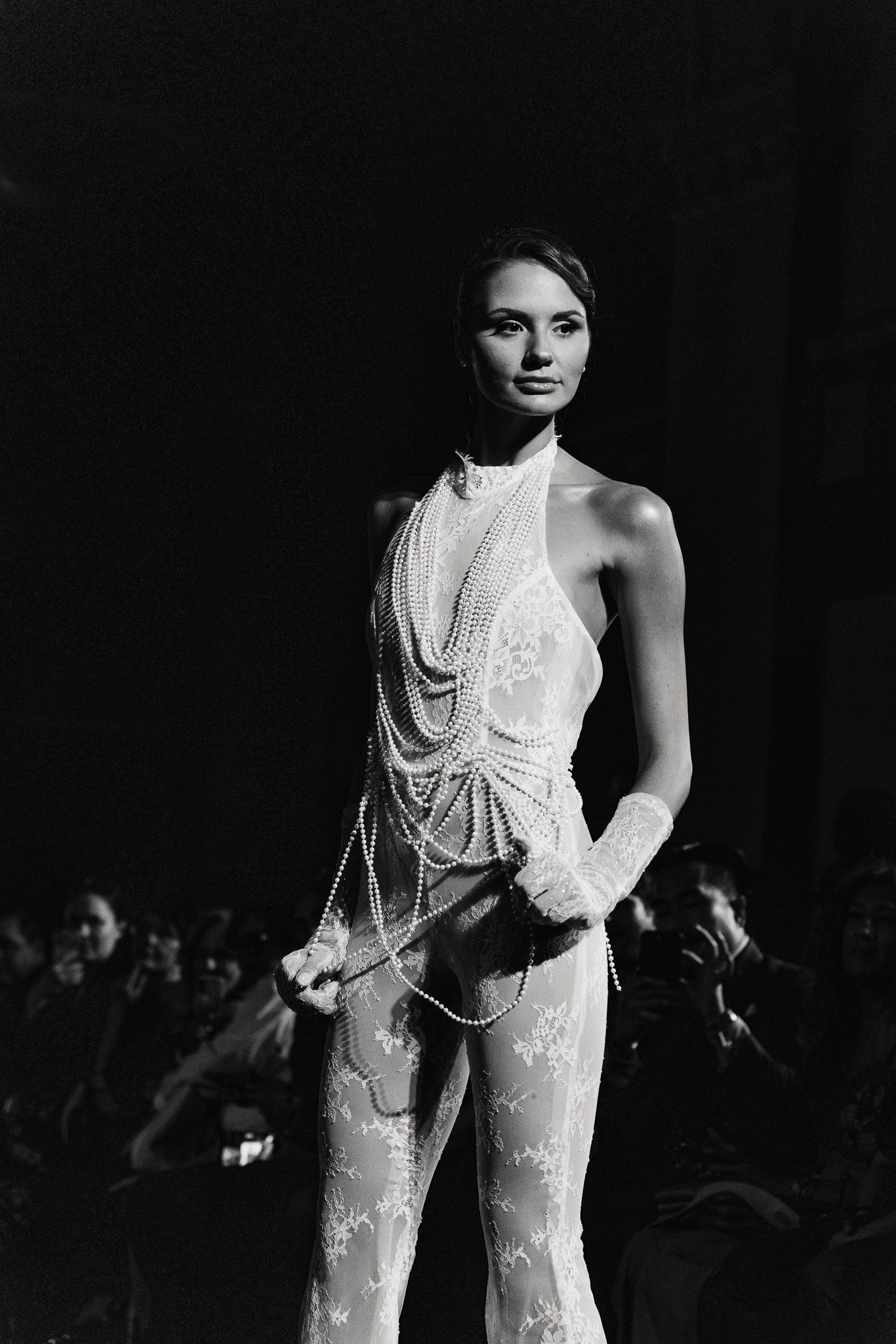 Black and white fashion photograph showing a model wearing a sheer white dress with pearl details against dark backdrop.