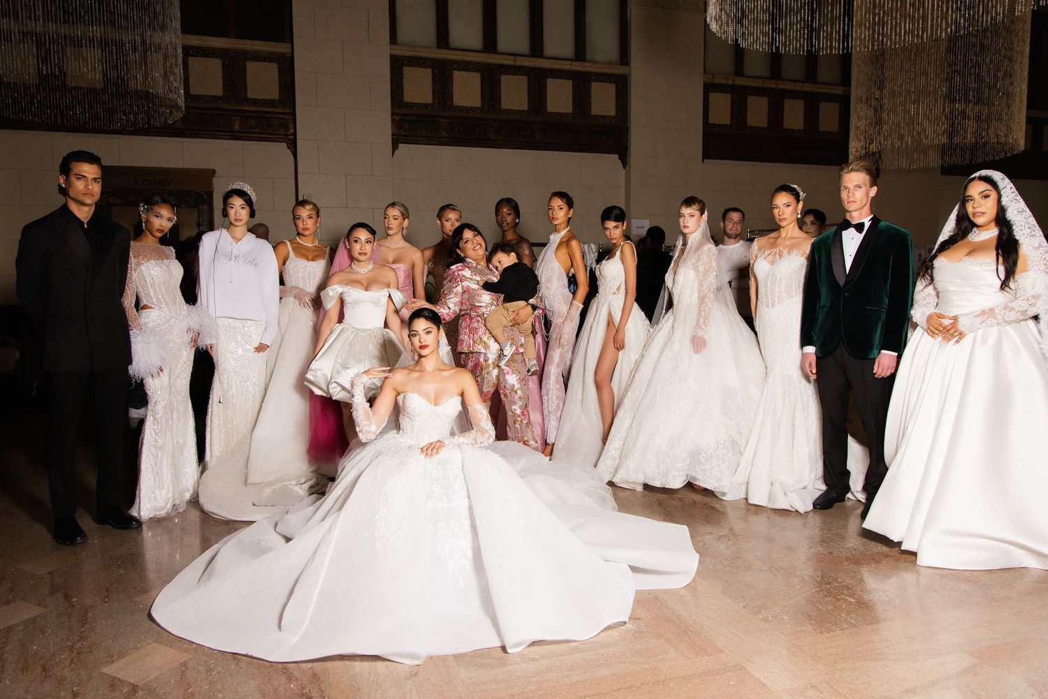 Group photo showing multiple people in white wedding dresses lined up in an indoor venue.