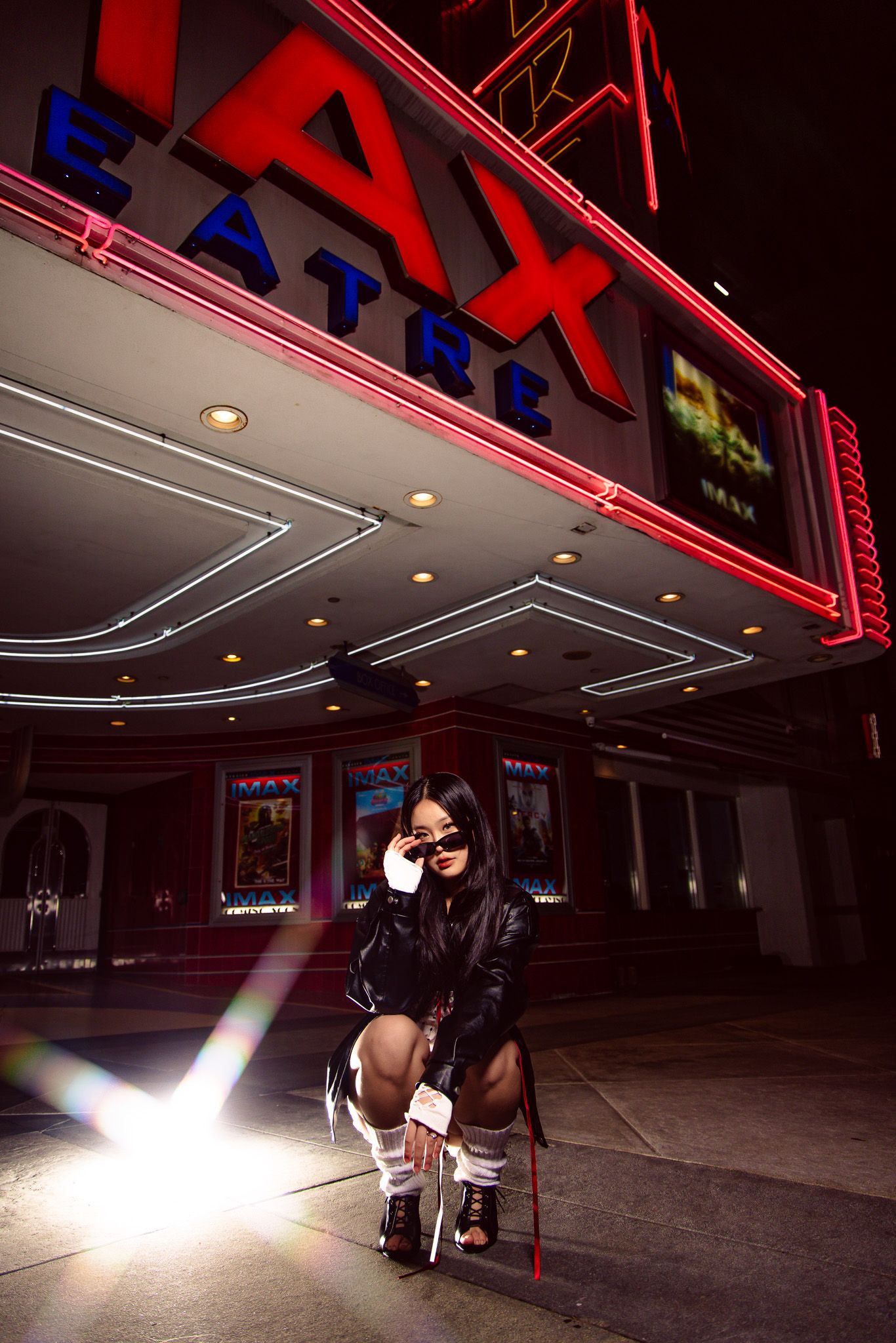 A dramatic nighttime scene outside a movie theater with neon signs and a bright light beam illuminating the entrance.