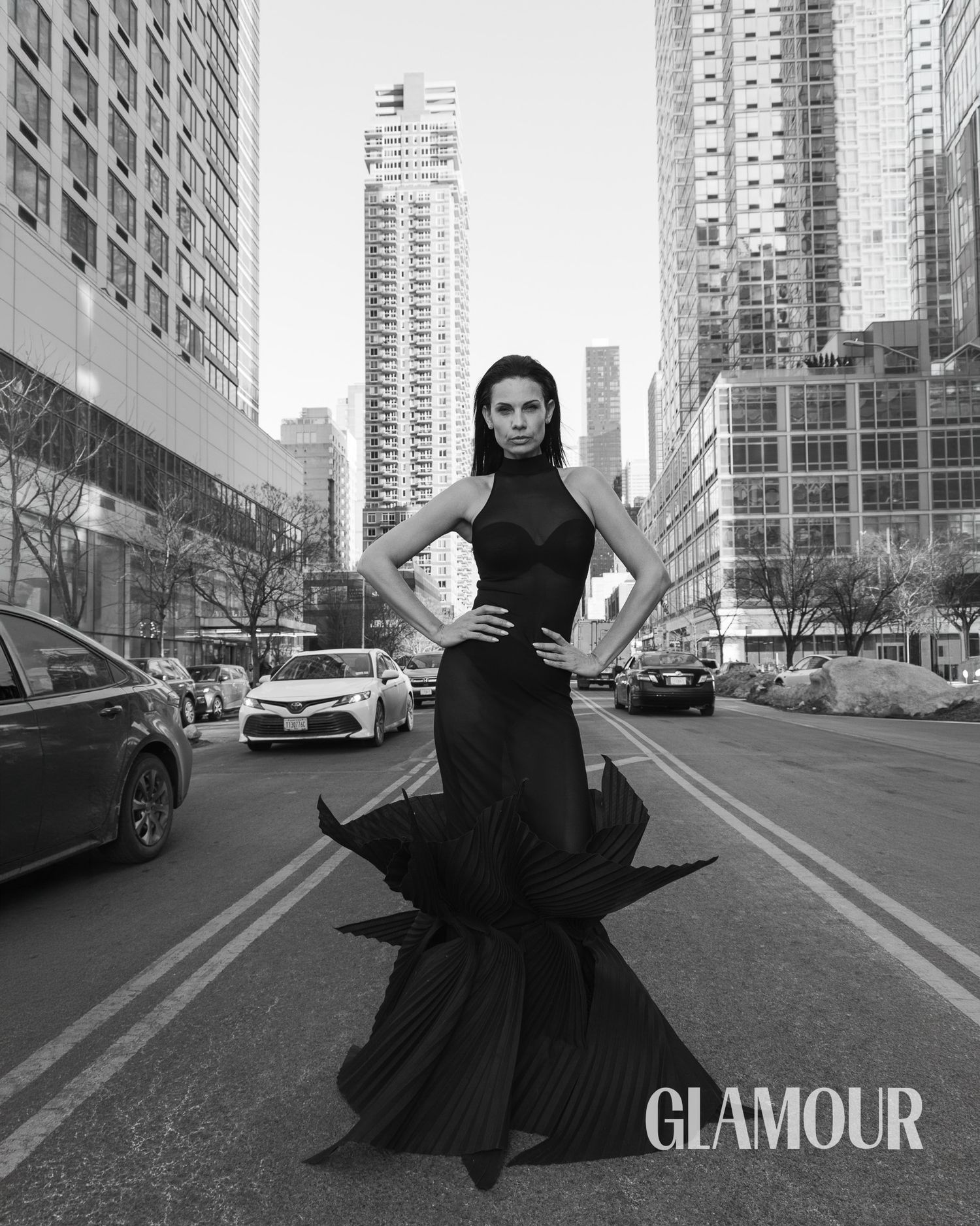 A black and white fashion editorial shows a model in dark clothing posing dramatically on a city street with tall buildings.