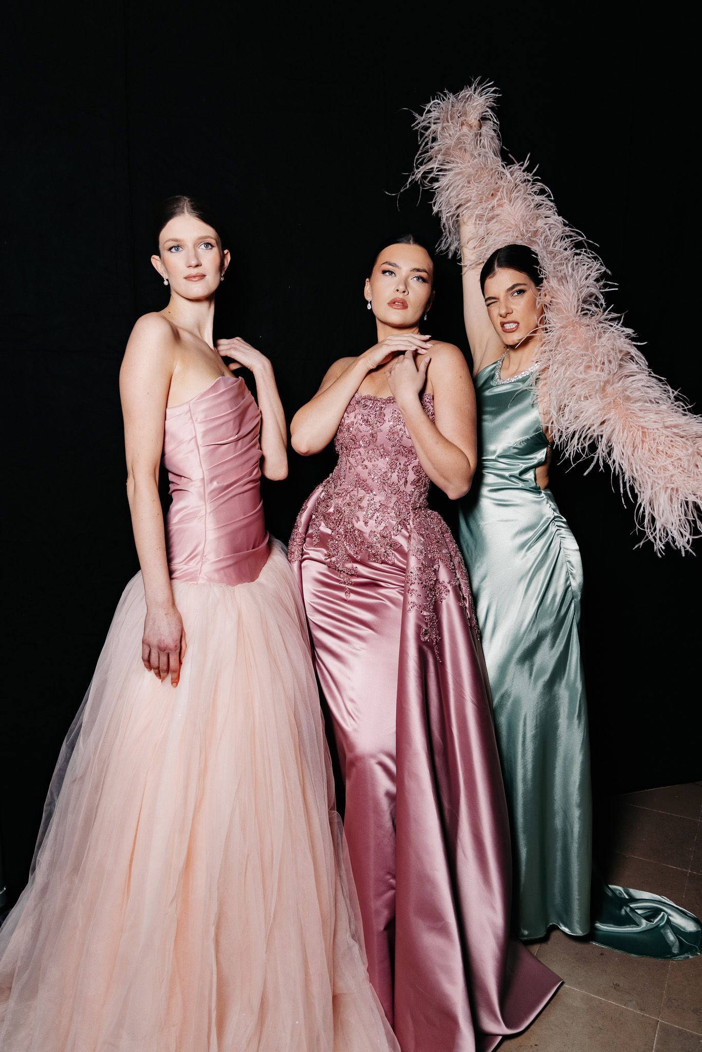 Three models pose in luxurious evening gowns featuring pink, rose, and mint satin with feather accents.