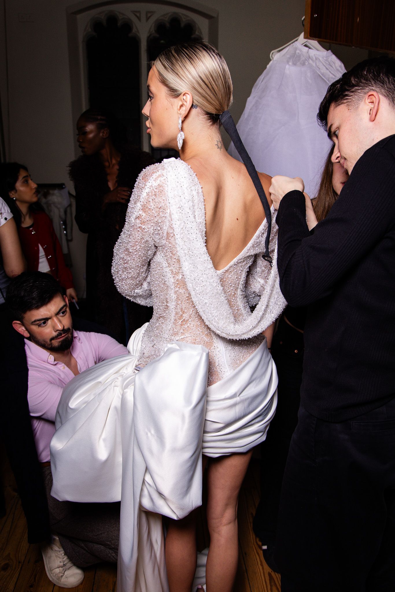 Behind the scenes at fashion event showing models in white dresses mingling with guests.