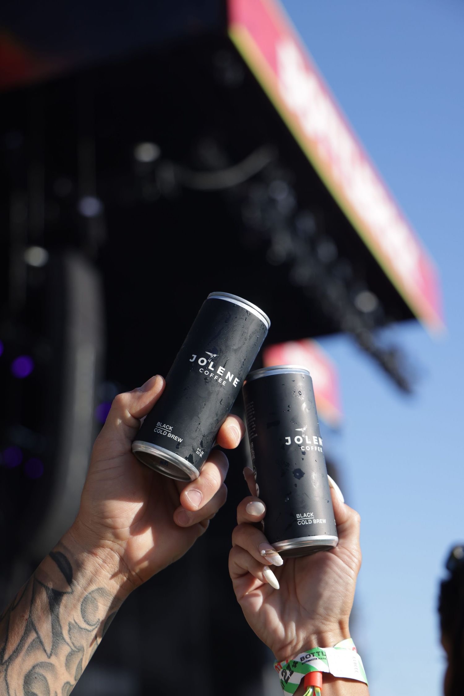Hands holding black beverage cans against a bright blue sky background.