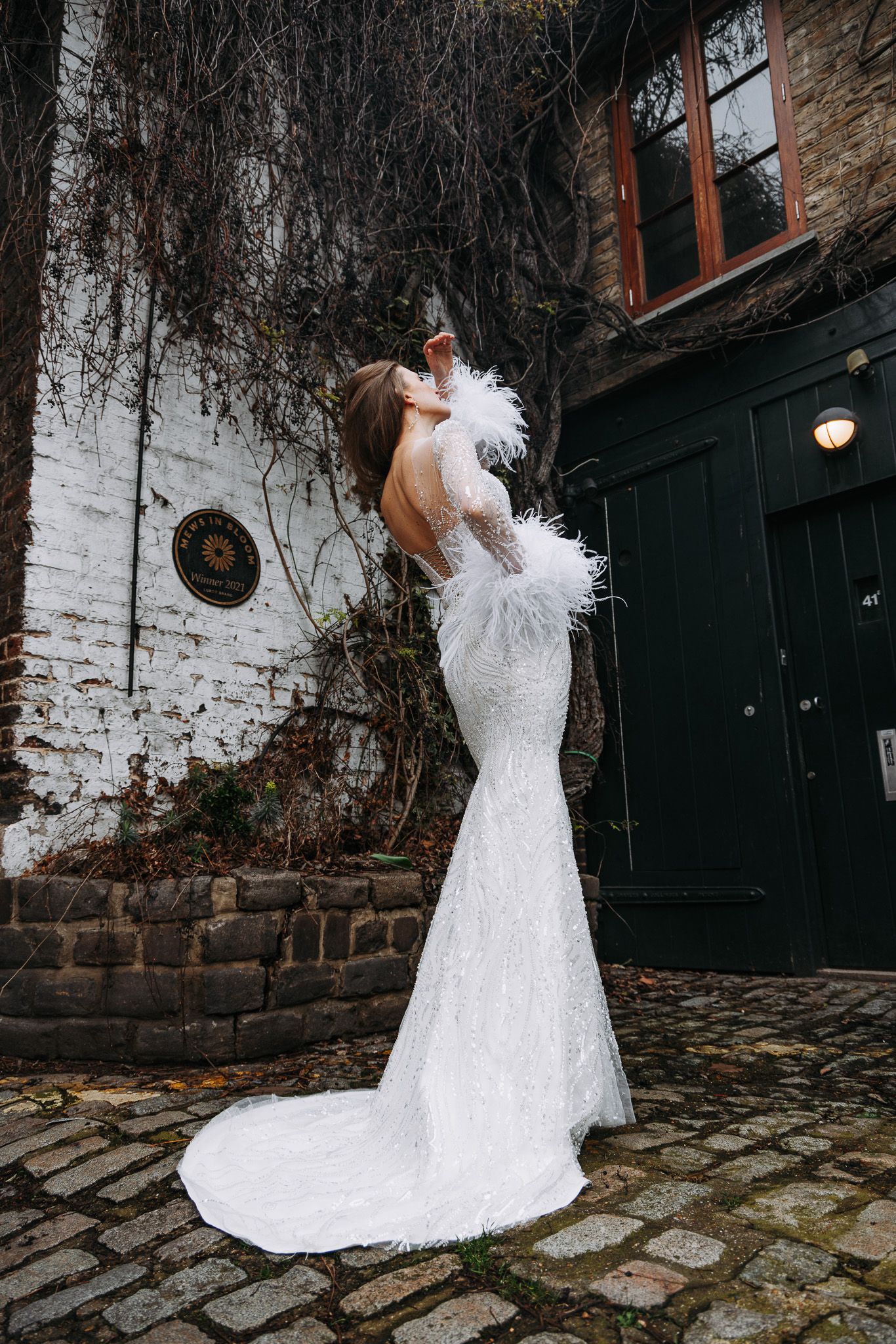 Elegant white mermaid wedding dress with feather details and long train photographed against rustic brick wall and cobblestone.
