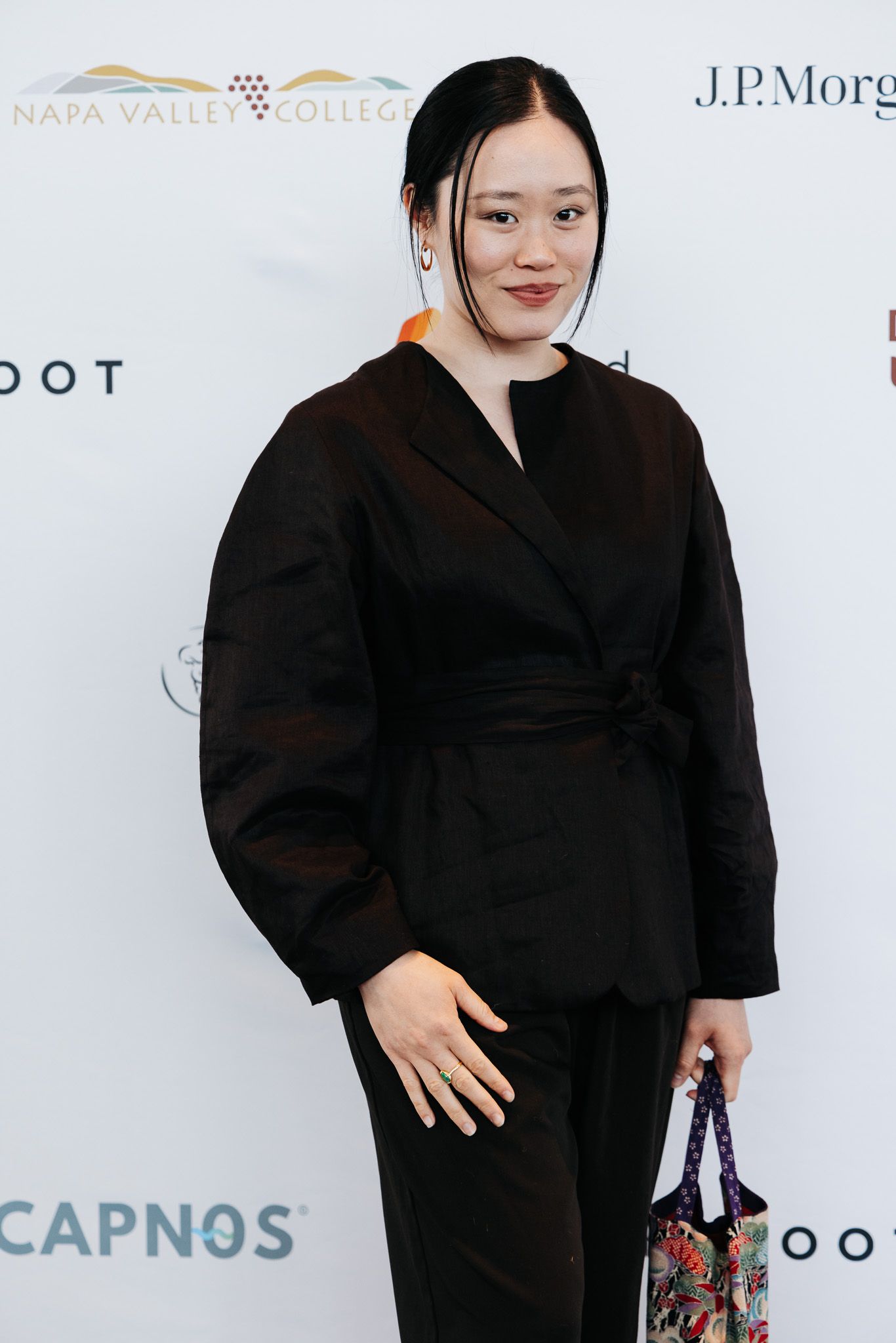 A person in an elegant black wrap dress with puff sleeves poses at an event with sponsor logos visible in the background.