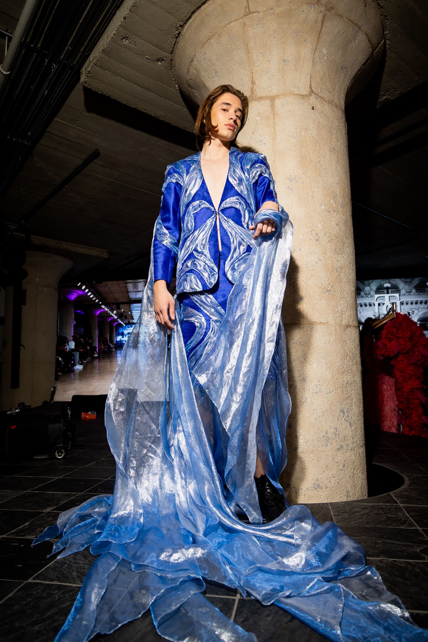 Model poses in flowing blue metallic dress against industrial concrete pillar backdrop in dramatic fashion photoshoot.